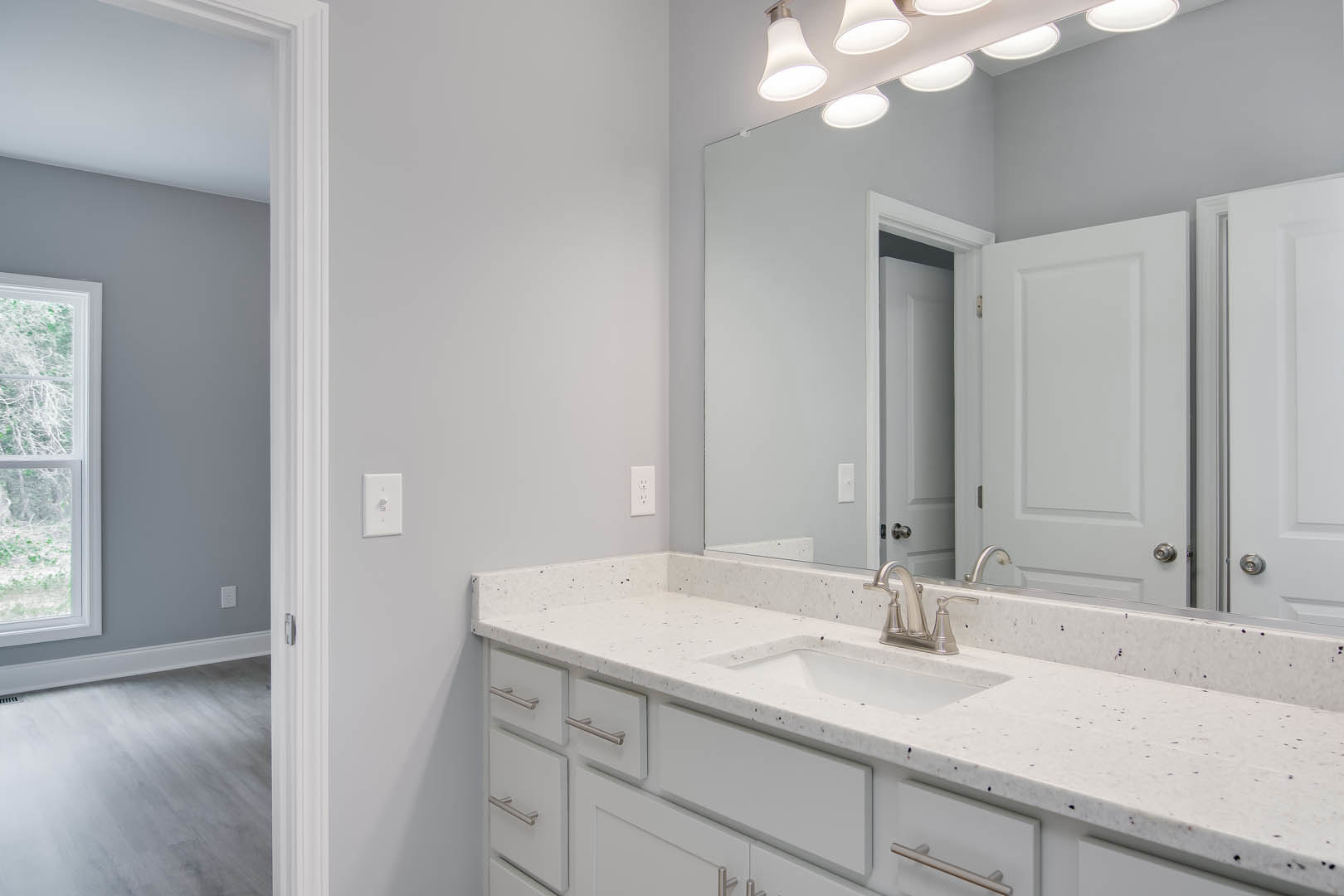 White bathroom with rectangular mirror above a wide sink, white countertop and cabinetry, chrome faucet, white framed window, light switch, white door, and wood floor with vent.
