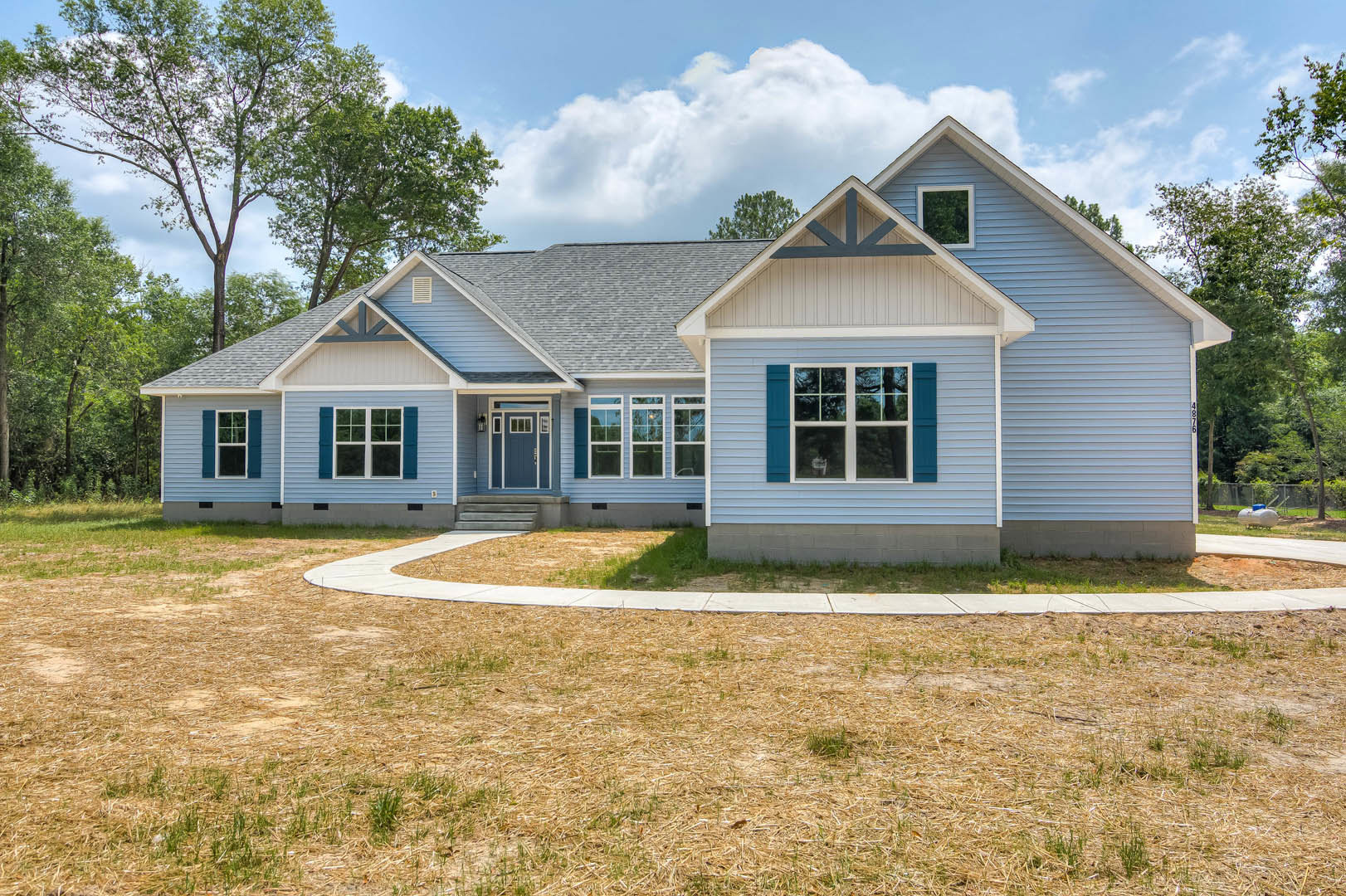 Two-story home with blue front door and white trim, large windows, grass lawn, paved driveway, and mature trees in the background