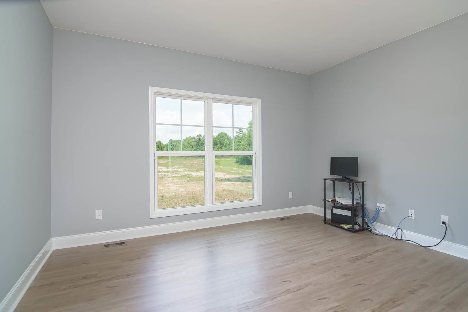 Living room with hardwood flooring, large window overlooking a grassy field, white walls, mounted television, floating shelf with a white storage box, and a computer monitor on a