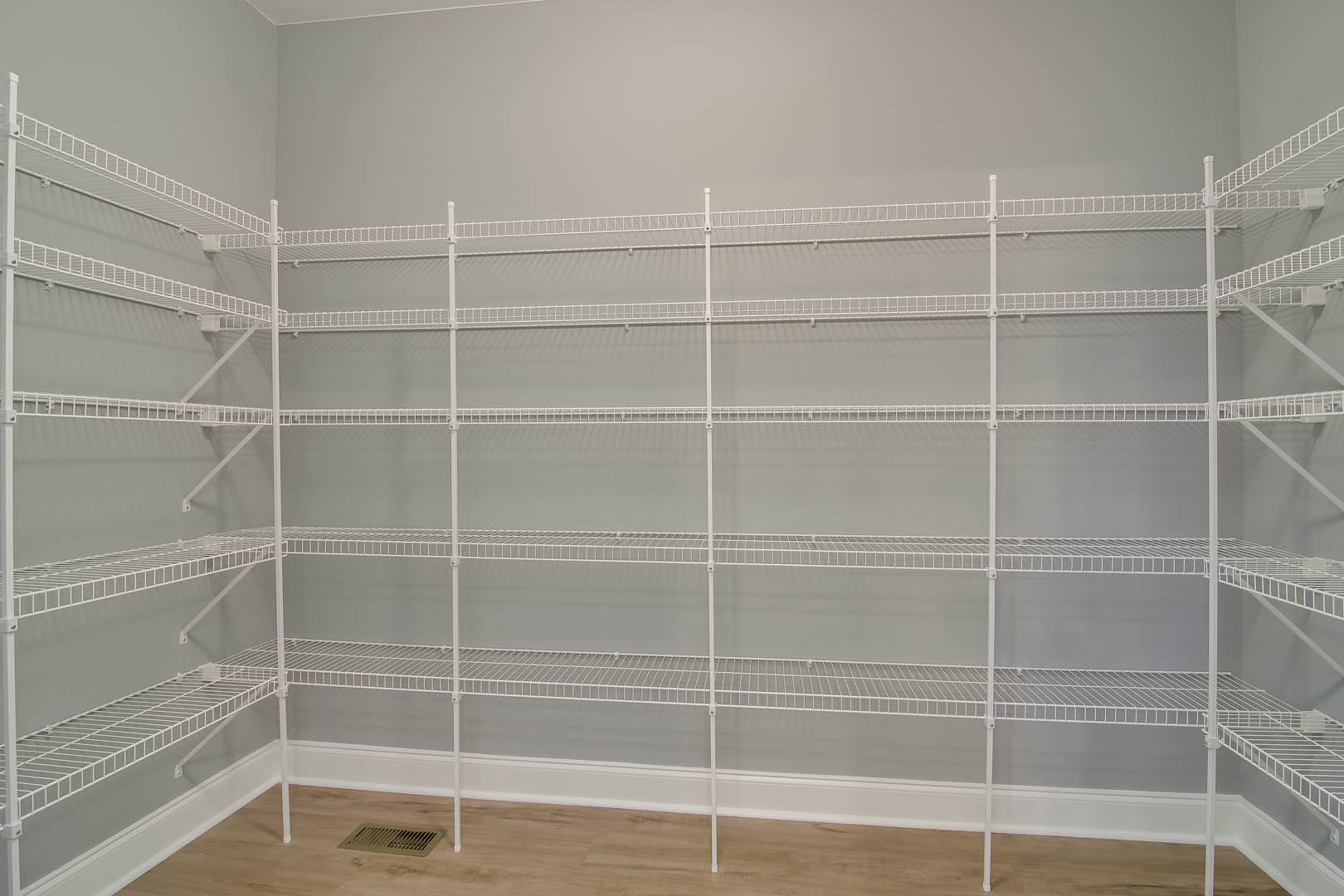 White wire shelving mounted on a wall in a closet, white metal supports, wood flooring, wall vent visible.