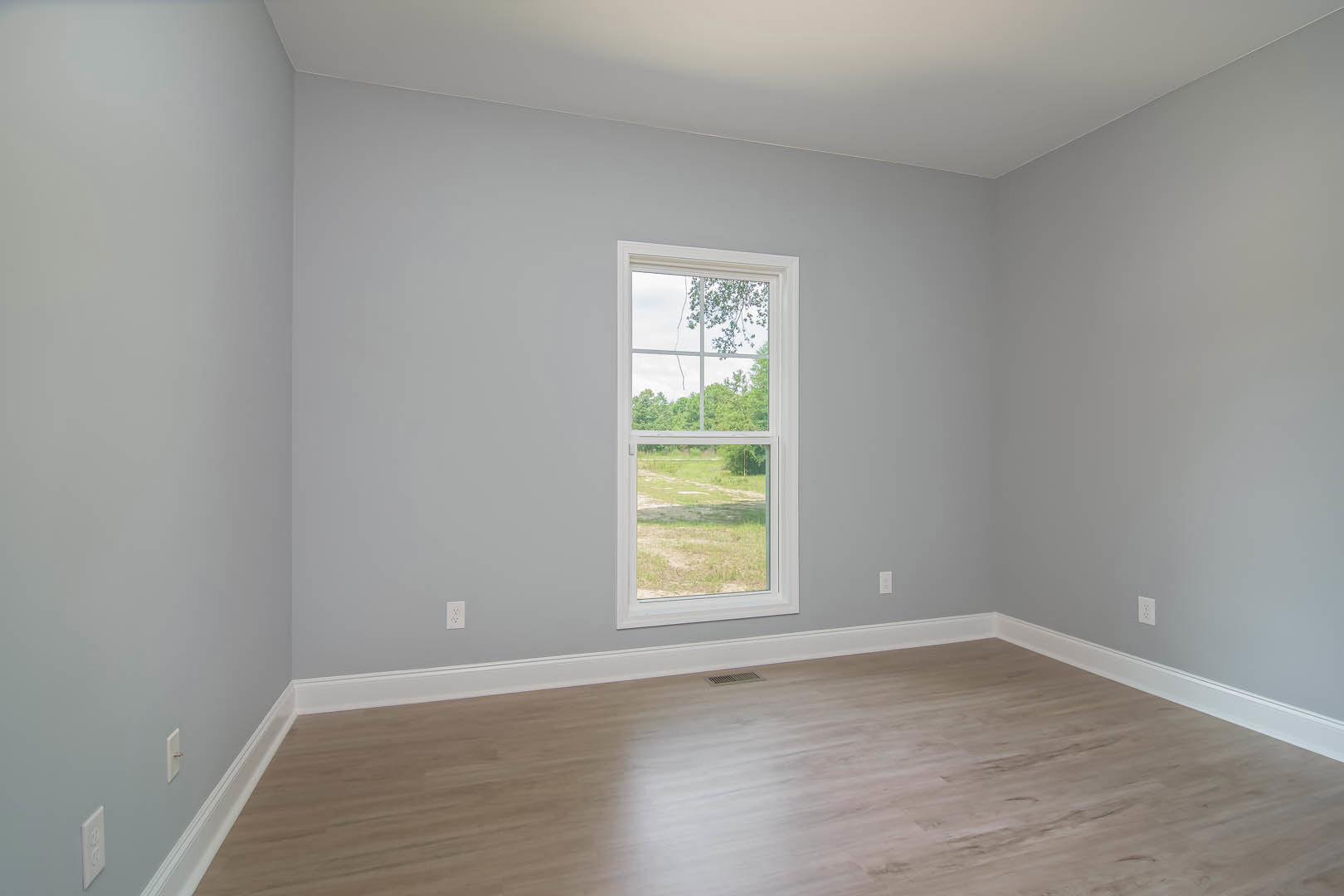 Sunlit room featuring wide-plank hardwood flooring, white walls, and a large window overlooking green trees and an open field; ceiling includes recessed lighting.