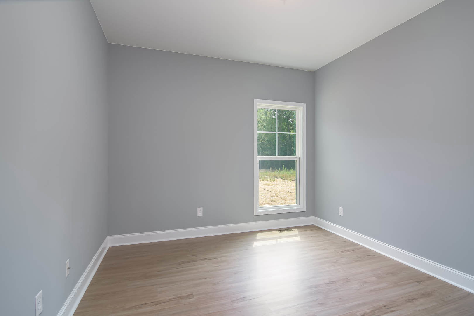 Sunlit room with white walls, white ceiling, and hardwood floor, featuring a window with a white frame overlooking a forested landscape.