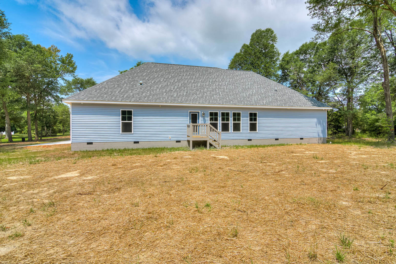 White farmhouse with wide front porch and wooden deck overlooking expansive grassy yard, surrounded by mature trees under partly cloudy sky