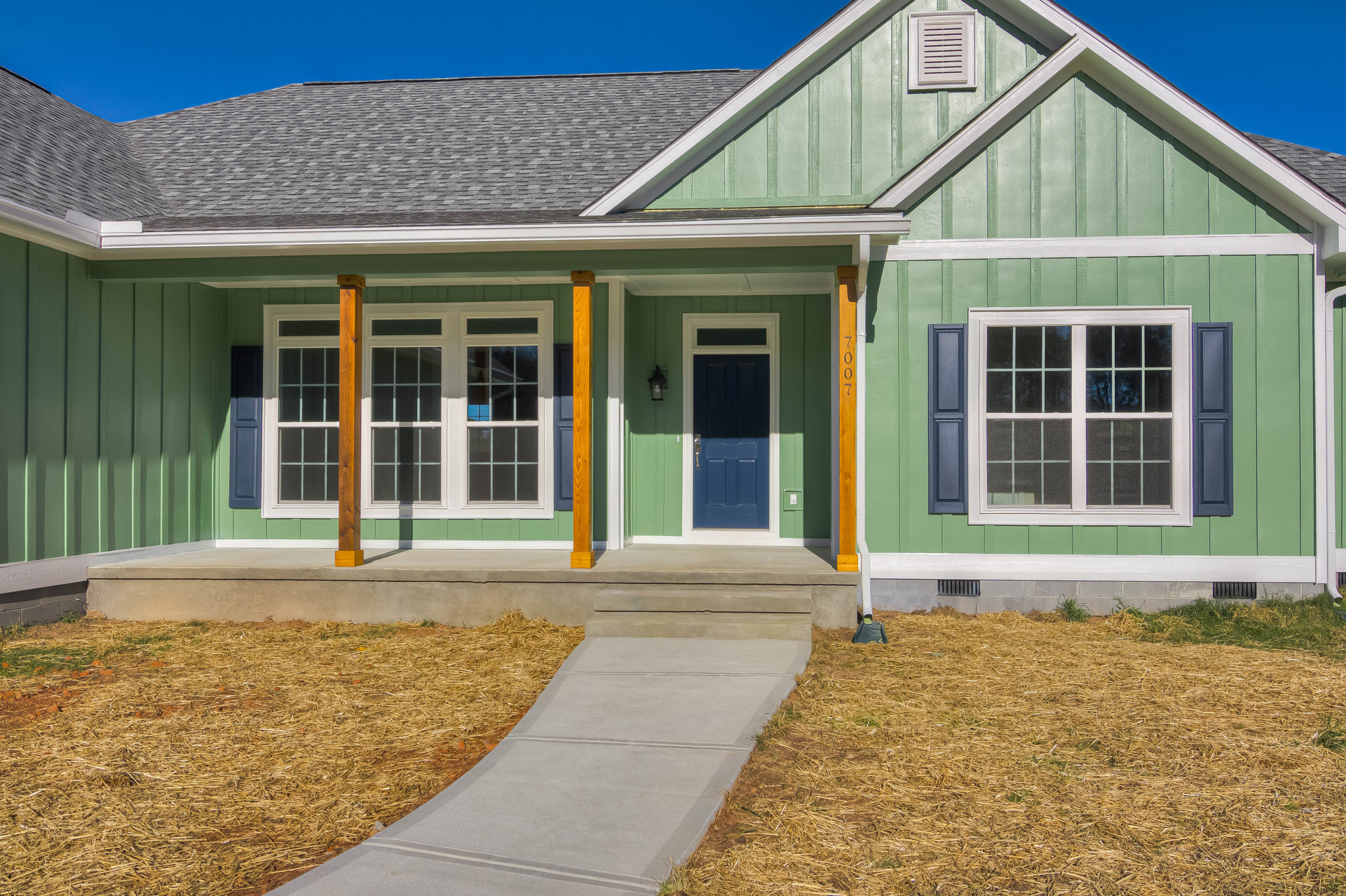 Concrete walkway leading to blue front door with white trim, white-framed window, gray siding, gabled roof, straw mulch along path, white vent below window