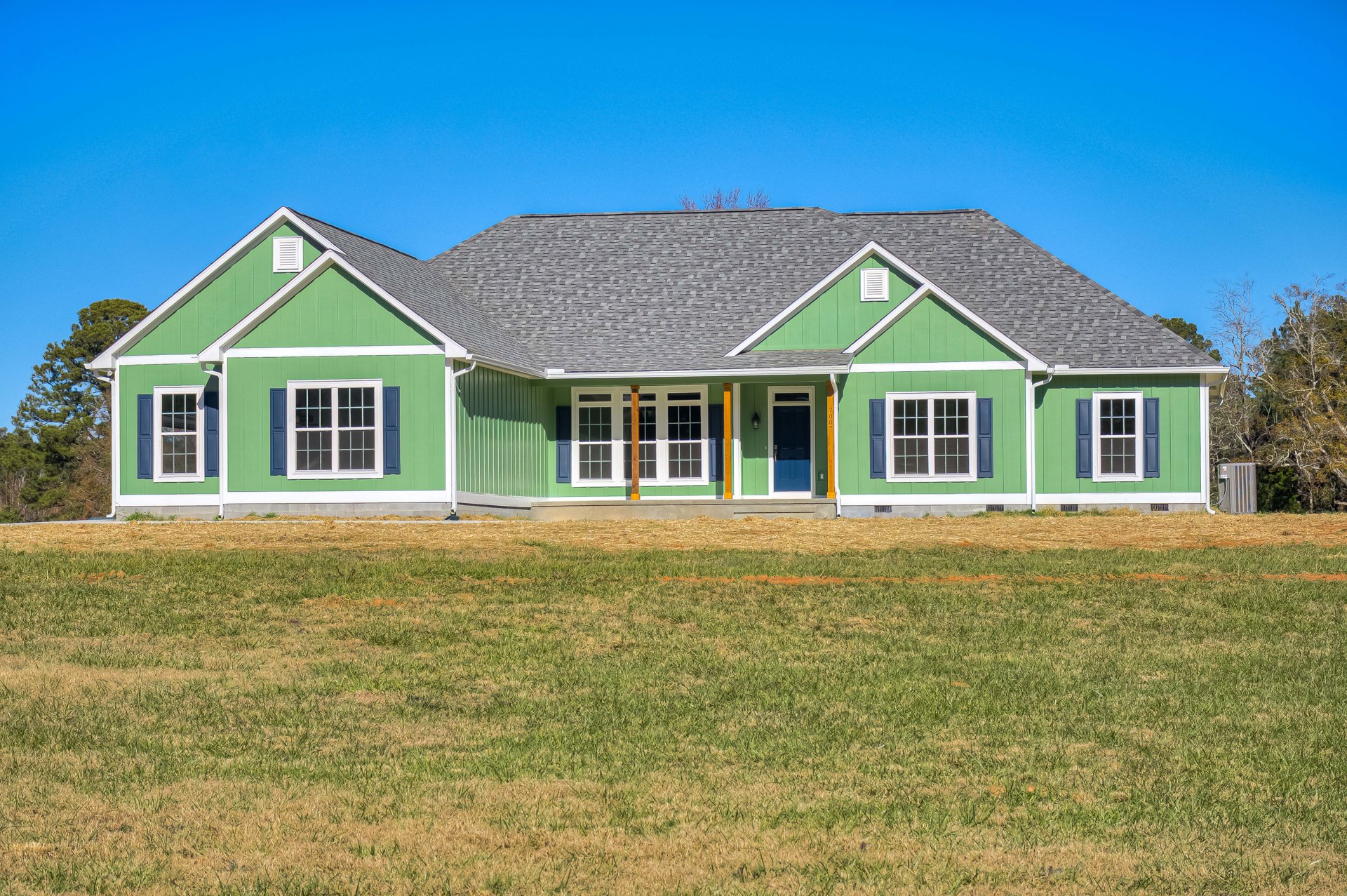 Green house with blue doors, white-framed windows, and blue shutters, surrounded by a large lawn with green and yellow grass under a clear sky