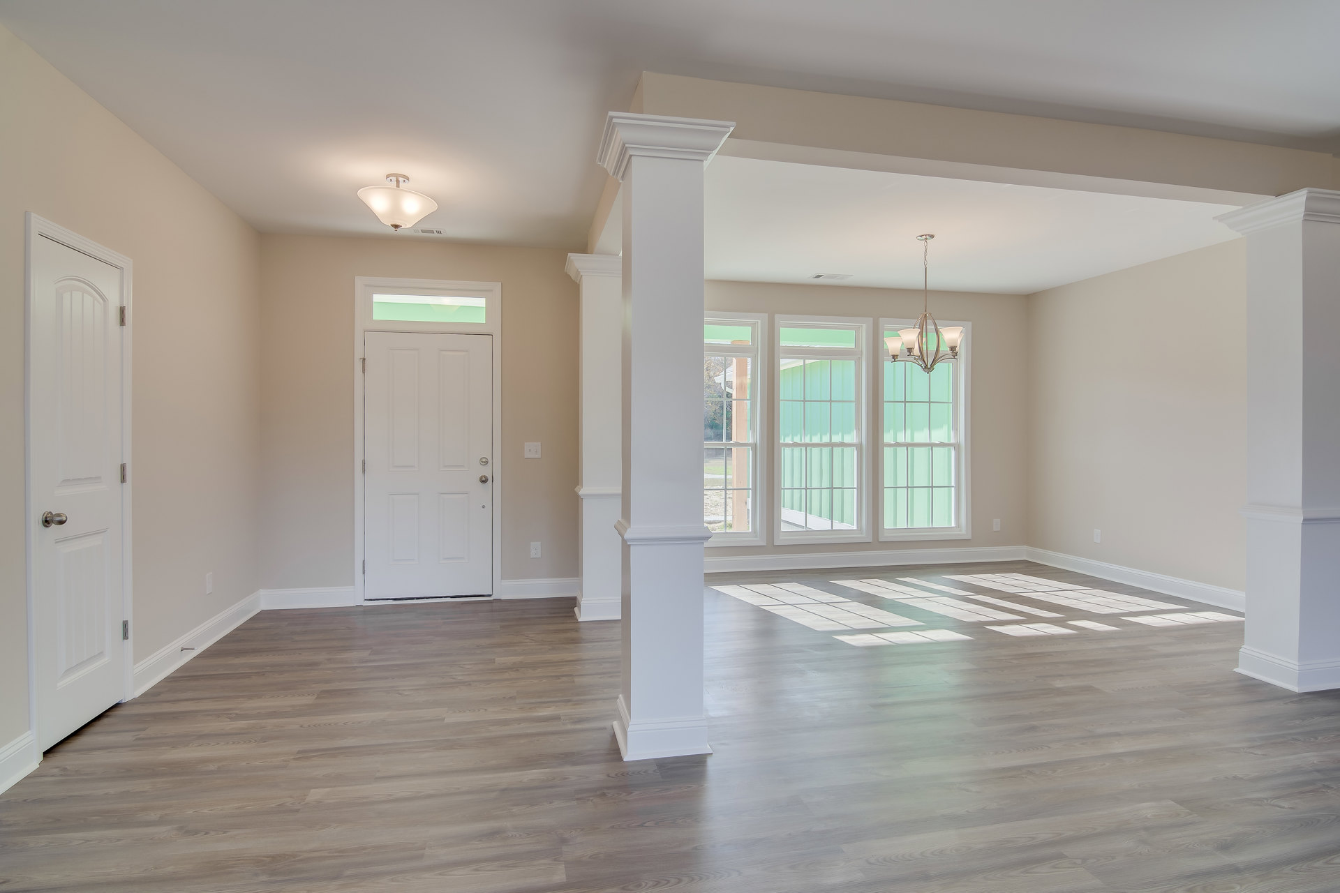 Wood flooring and white columns in a bright room with a white door featuring a silver knob, glass-paned window, and ceiling light fixture.