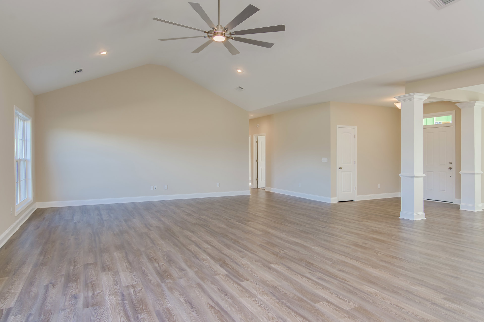 Spacious room with hardwood flooring, white and grey walls, ceiling fan with light fixture, white door with silver doorknob, and large window