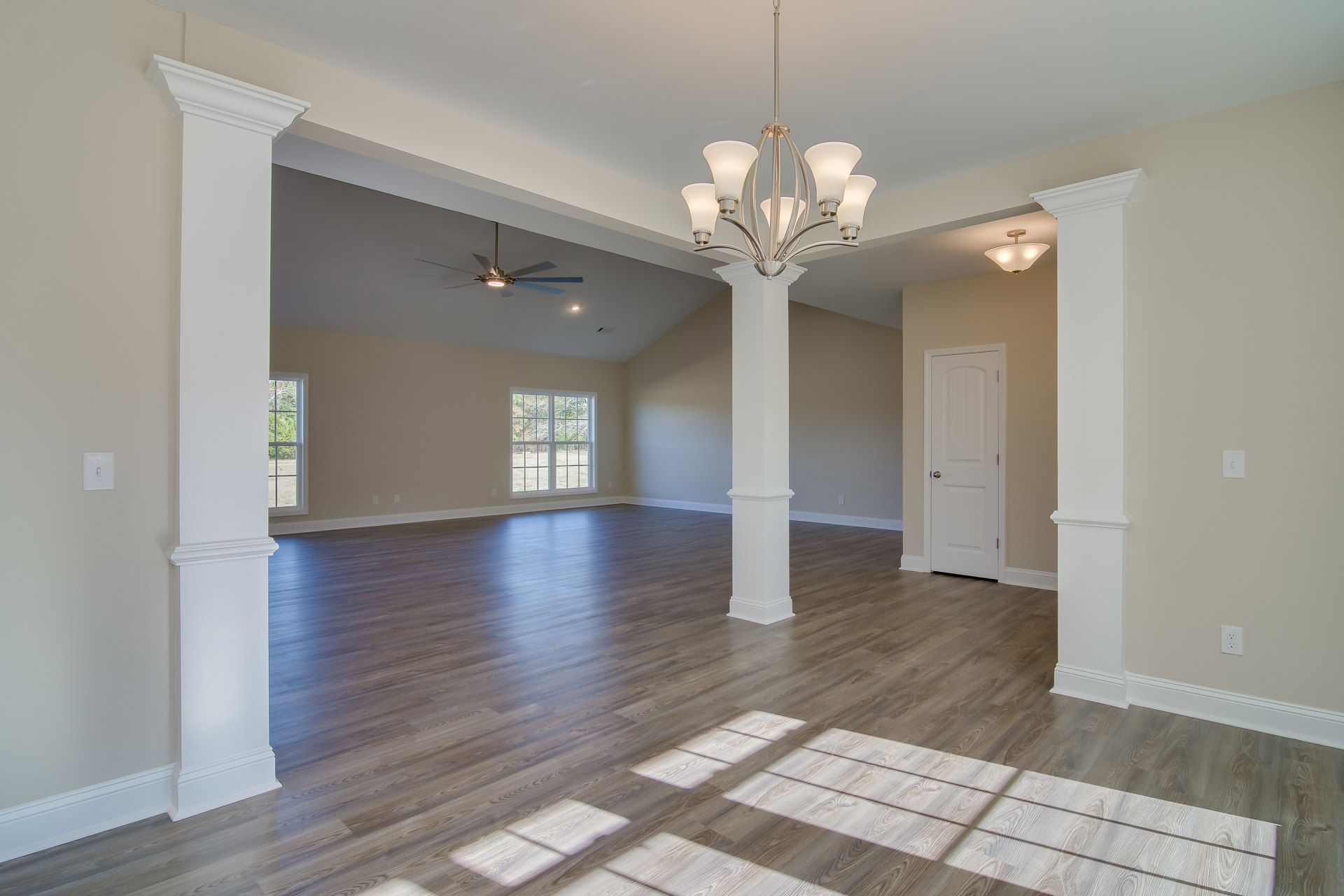 Spacious room with wood flooring, white pillars, large windows overlooking trees, white door with silver doorknob, and five-light chandelier hanging from ceiling