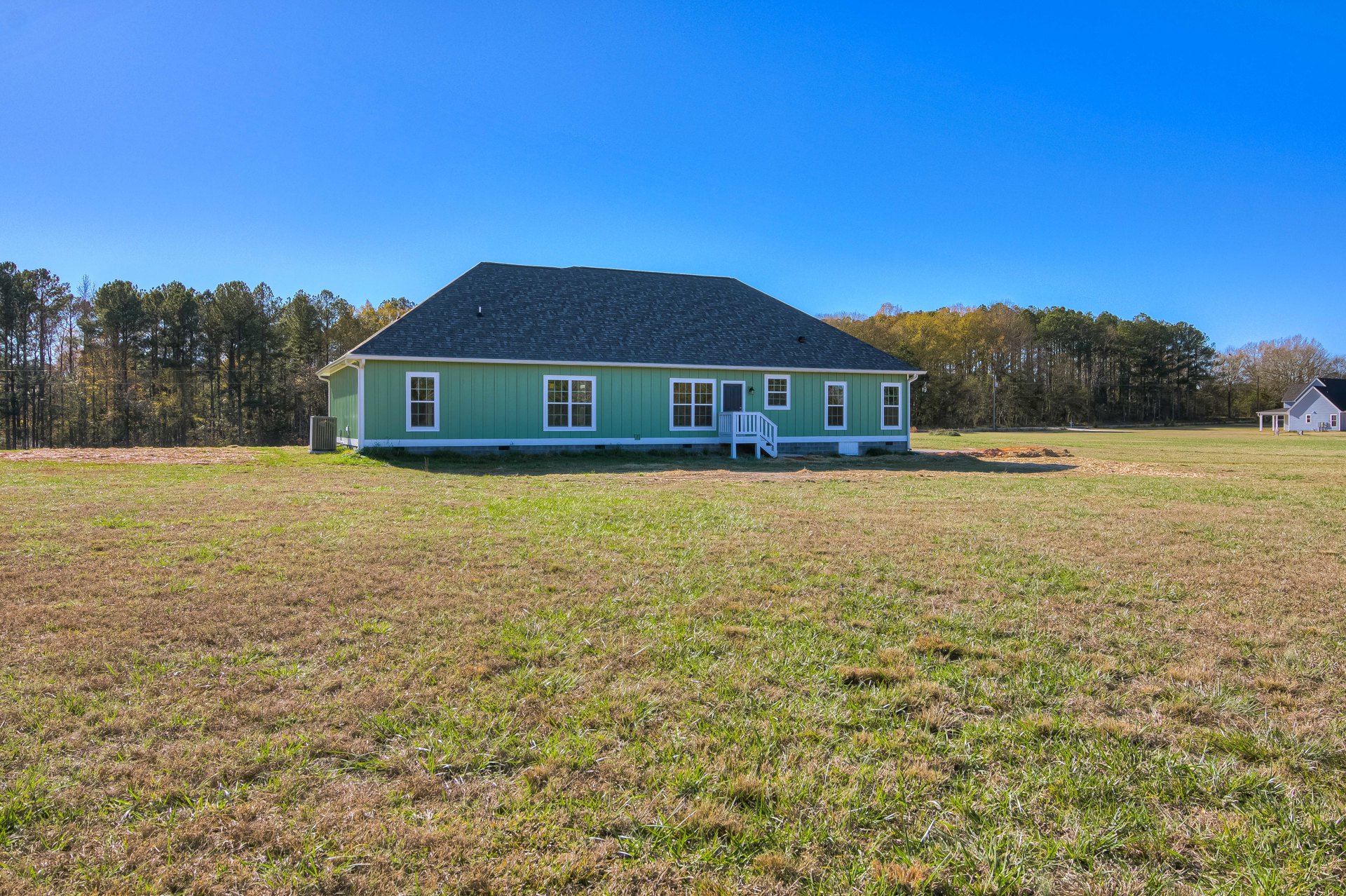 Green-painted cottage with white-trimmed windows, spacious porch featuring white railing, surrounded by a large grassy field and mature trees under a clear sky.