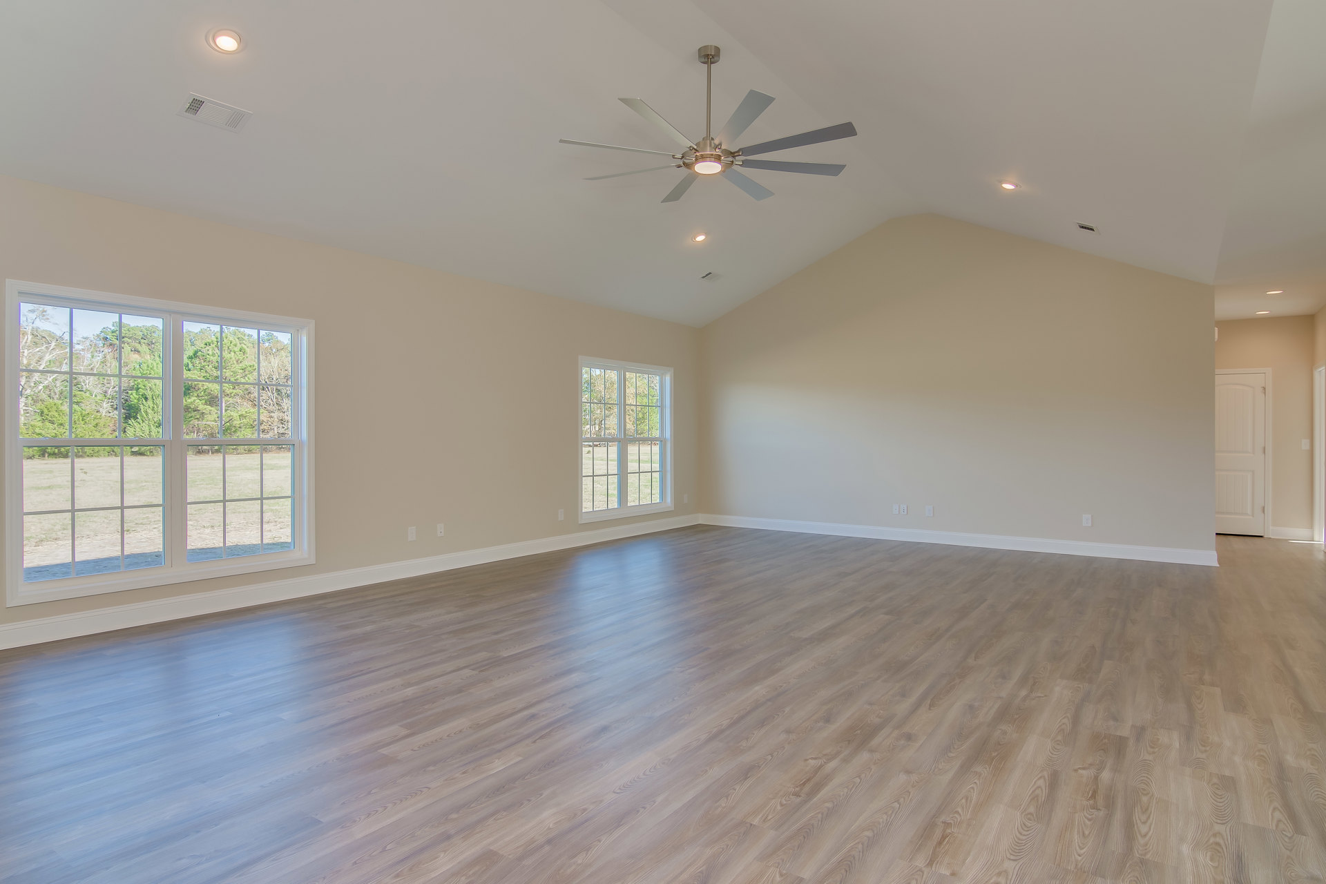 Spacious room with wood flooring, ceiling fan with light fixture, multiple windows showing leafy trees outside, and white doors against light-colored walls