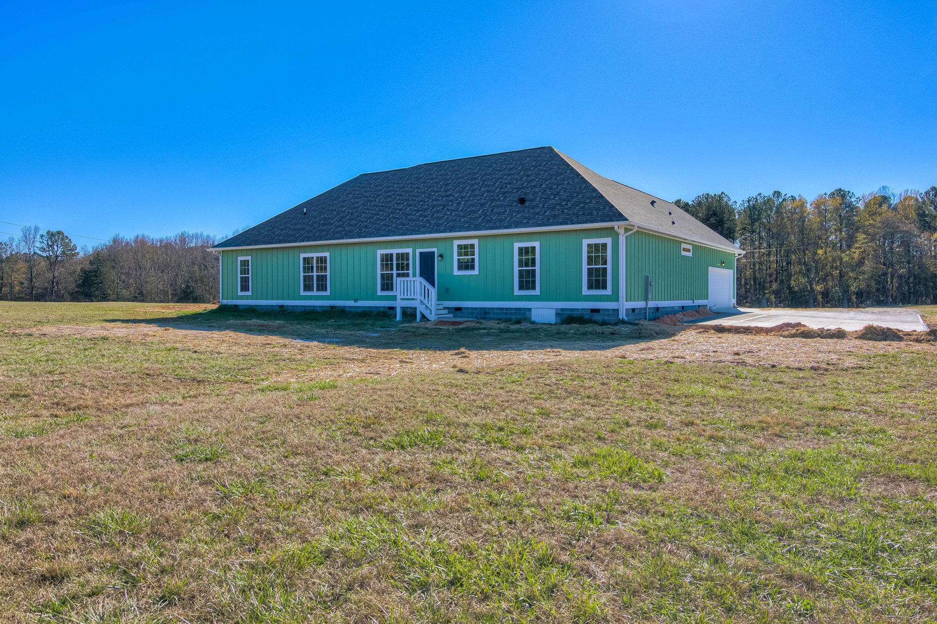 Green-painted house with white staircase, expansive grassy lawn, white porch, black front door, large windows, and blue sky above