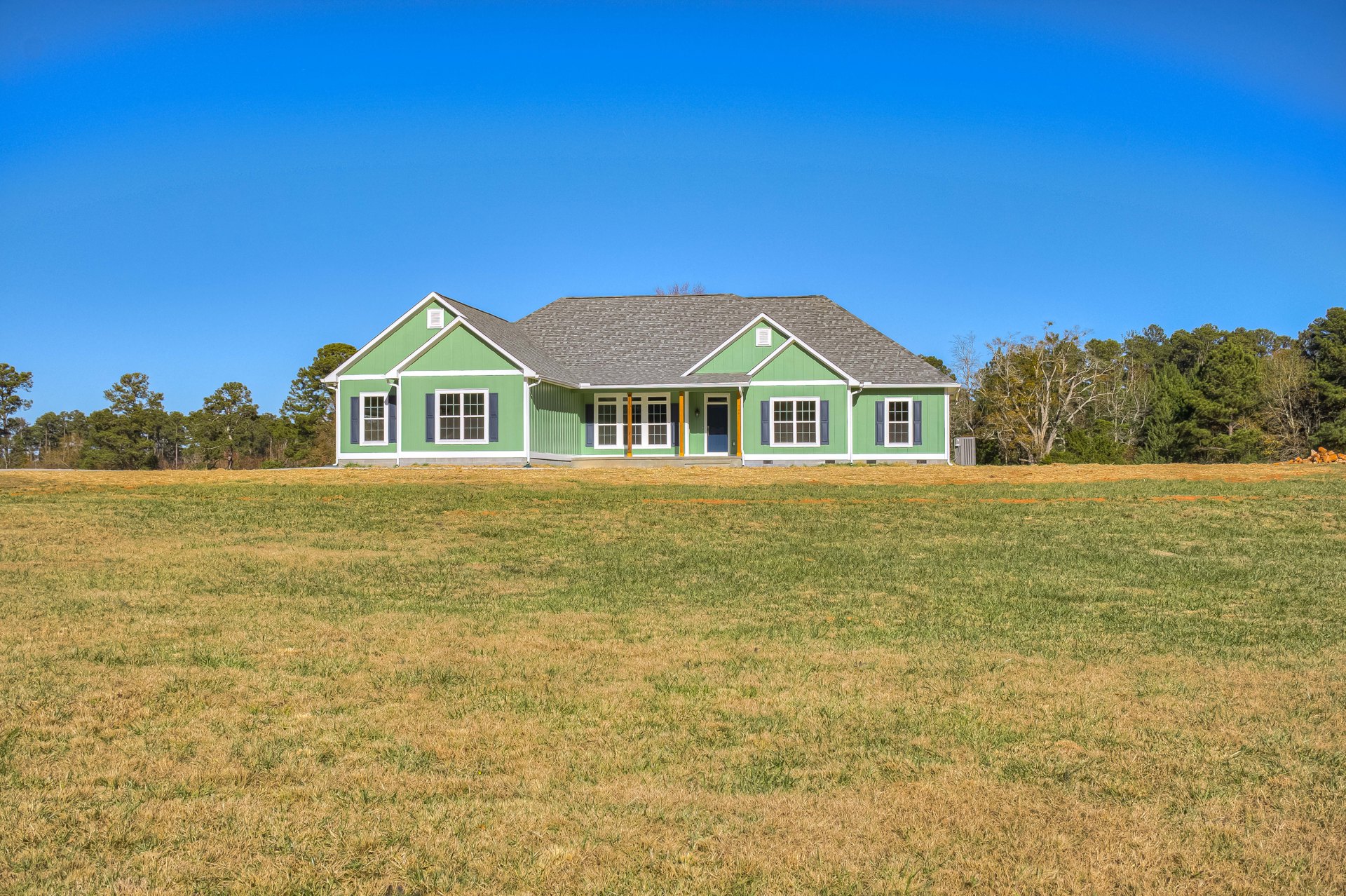 Green-painted house with white trim set on a spacious grassy field, surrounded by trees under a clear blue sky
