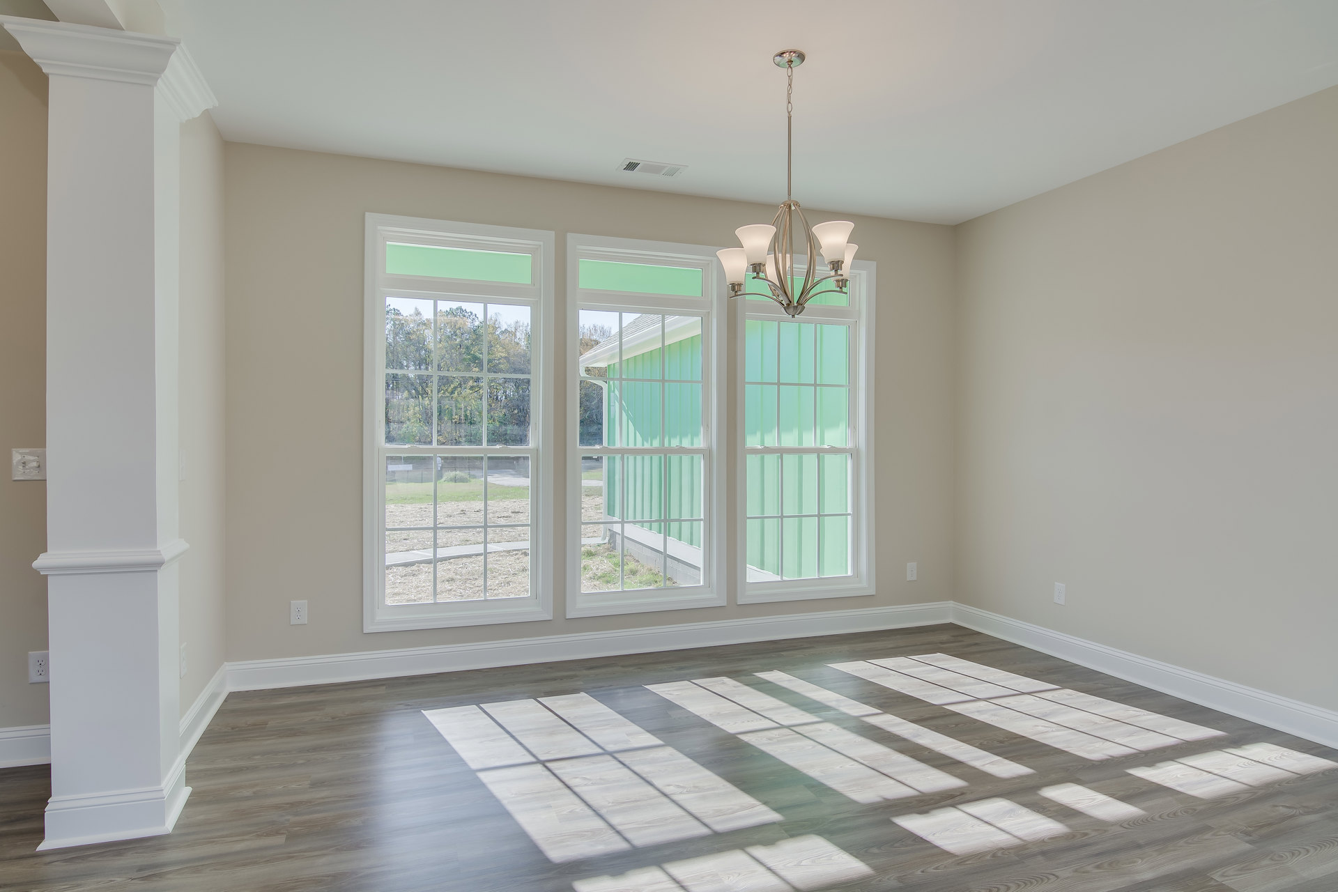 Spacious room with large windows, white and grey flooring, four-light chandelier hanging from ceiling, decorative wall molding, view of trees outside