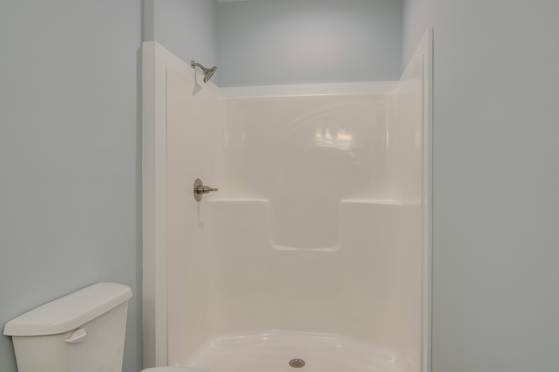 White tiled shower with chrome shower head next to a white toilet tank and handle in a bathroom.
