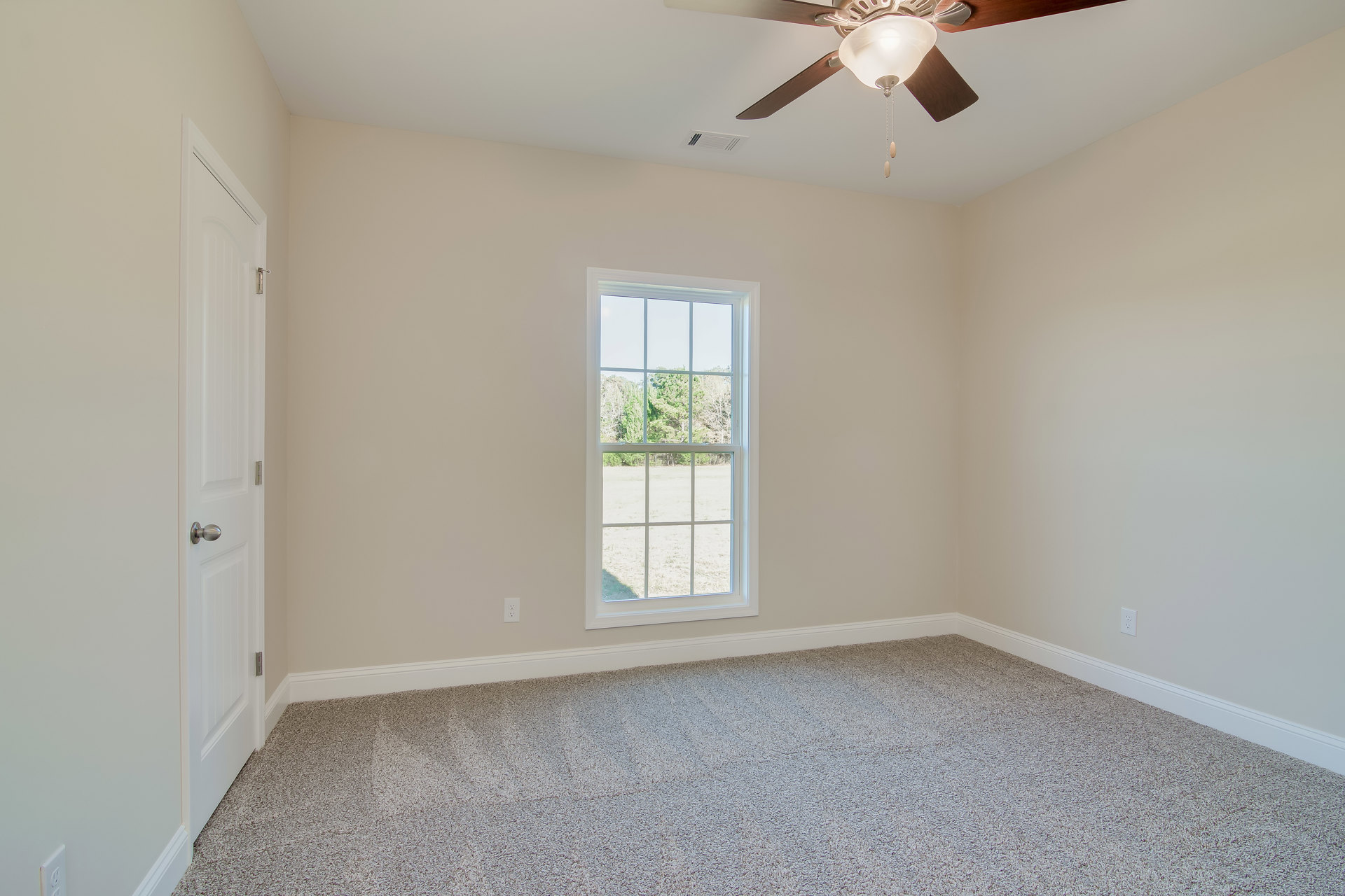 Carpeted room with white walls, ceiling fan and light fixture, large window showing trees outside, white door with silver knob, plaster ceiling with crown molding.