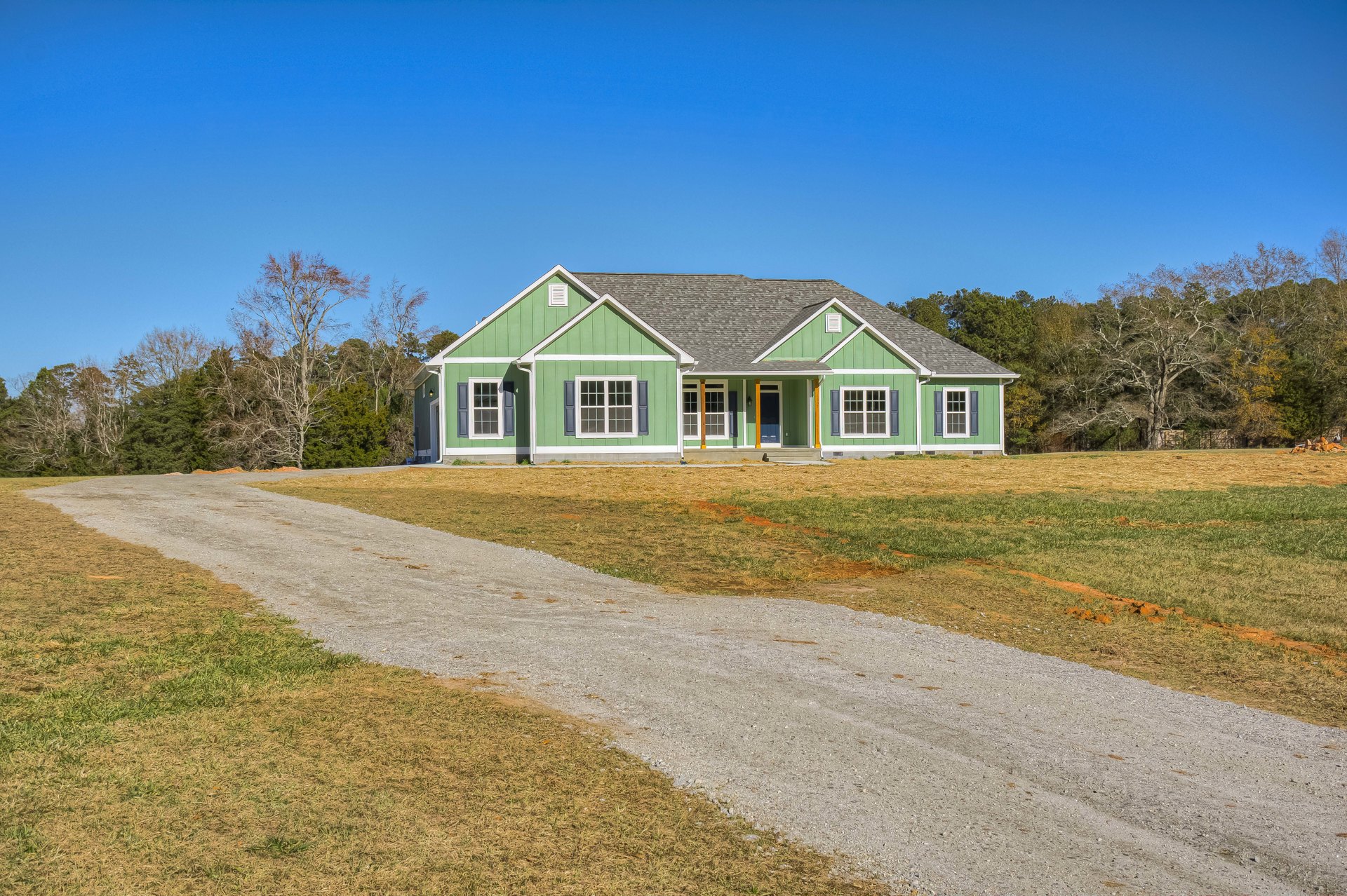 Green house with white trim, gravel road bordered by grass, leafless tree nearby, blue sky overhead, close-up window visible on facade