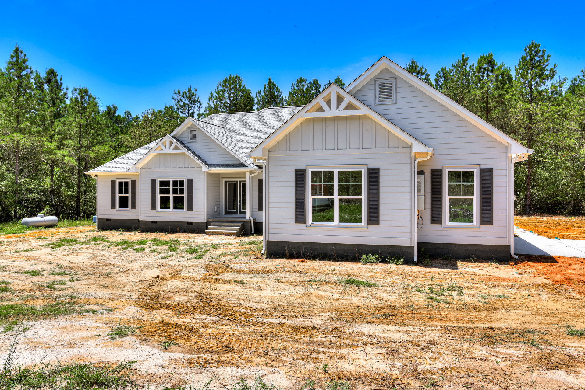 Partially built house with exposed framing, white window frames, wall vent, and door; dirt patch and bicycle on grass in front; mature trees in background.
