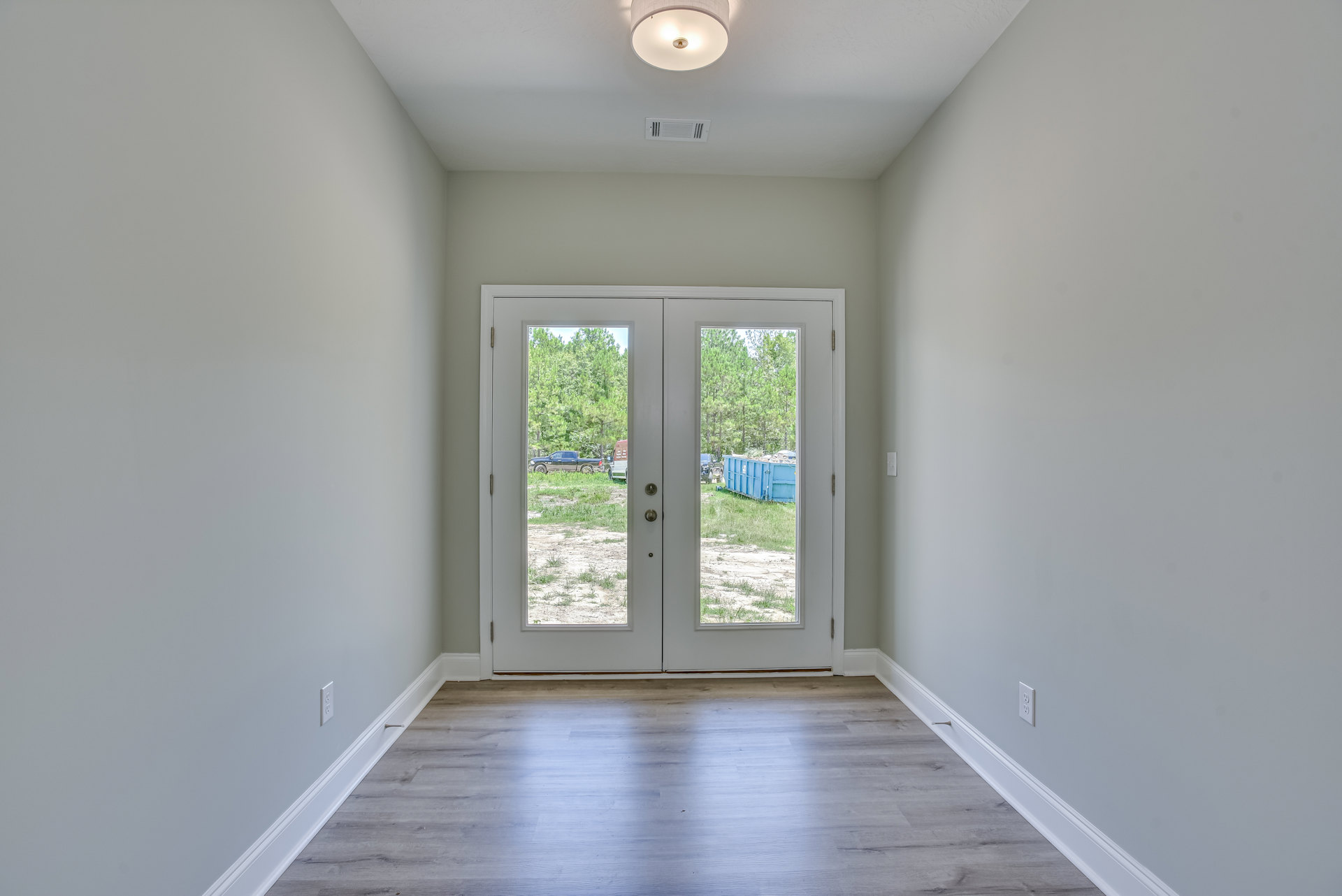 Hallway with wood flooring, double doors featuring glass panes, ceiling light fixture and vent, sunlight illuminating the floor, blue dumpster visible outside through the doors