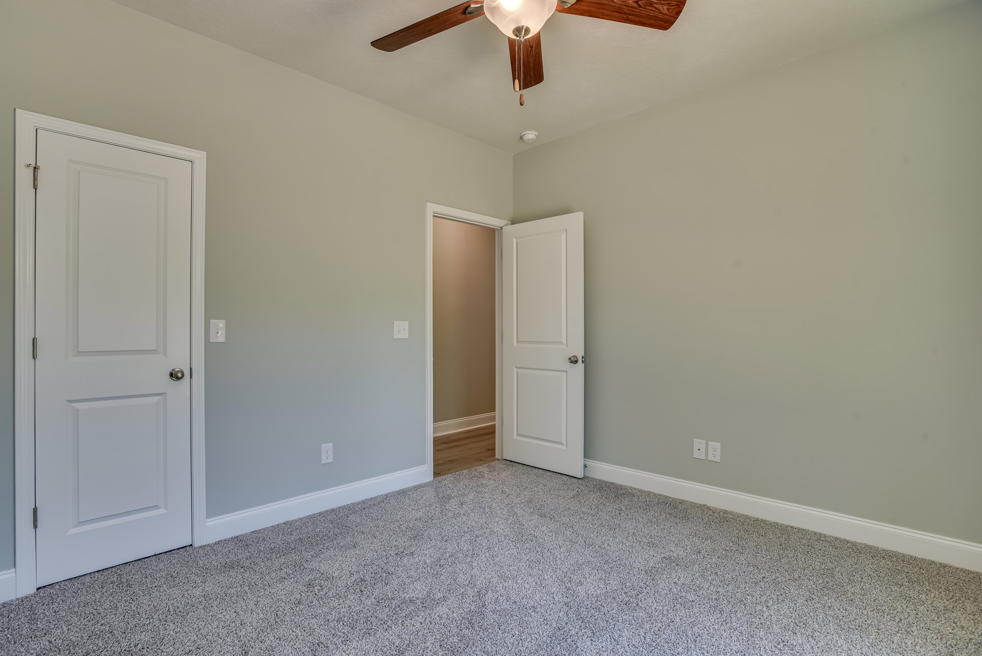 Neutral-toned room with brown walls, white trim, ceiling fan with light fixture, two white doors—one open with wood interior, one closed with silver knob, light-colored flooring.