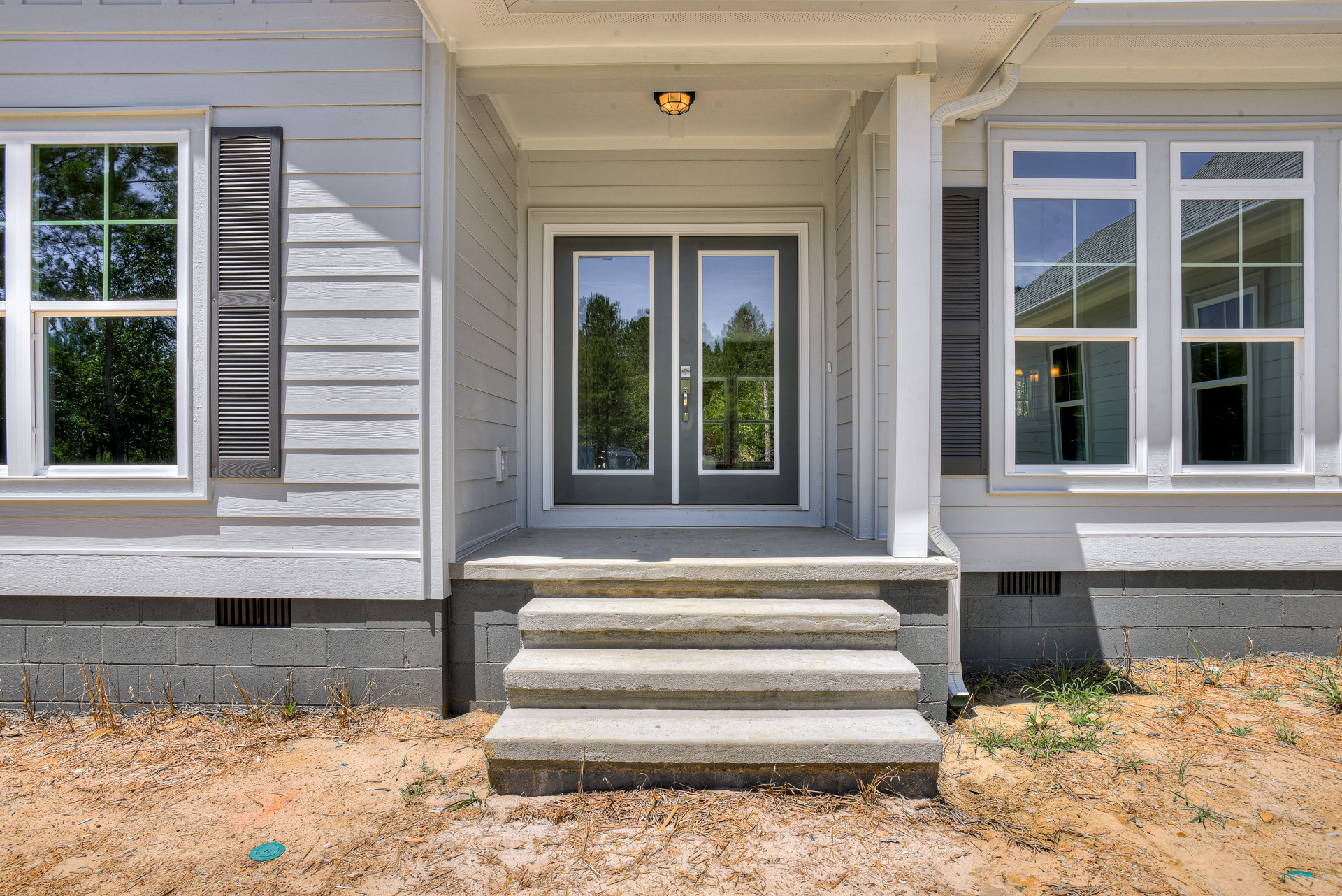 Wooden front door with glass panels, flanked by windows, set on a covered porch with stairs, surrounded by landscaped plants and trees