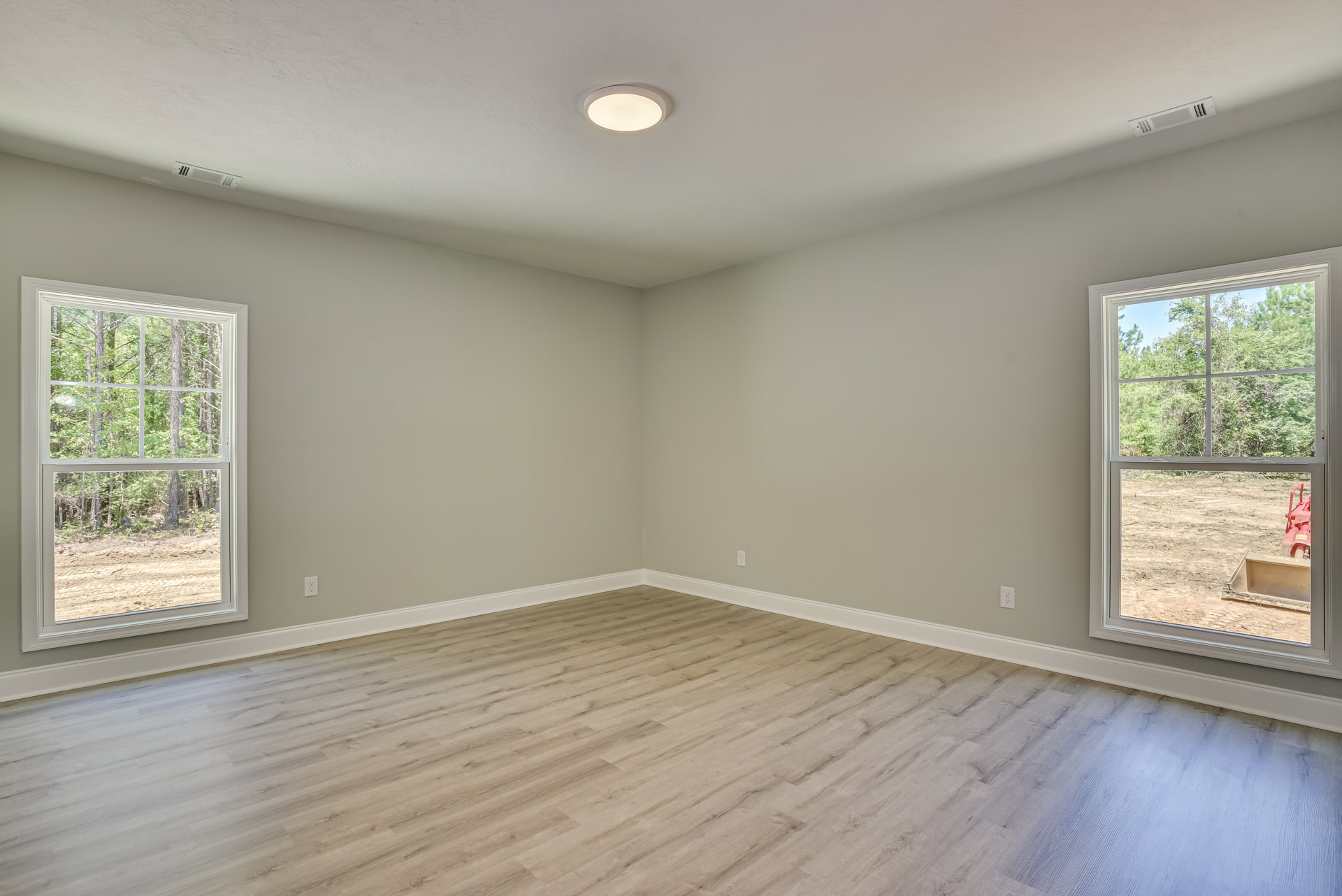 Wood flooring with white baseboards, ceiling light fixture, large window overlooking trees and a tractor in the dirt