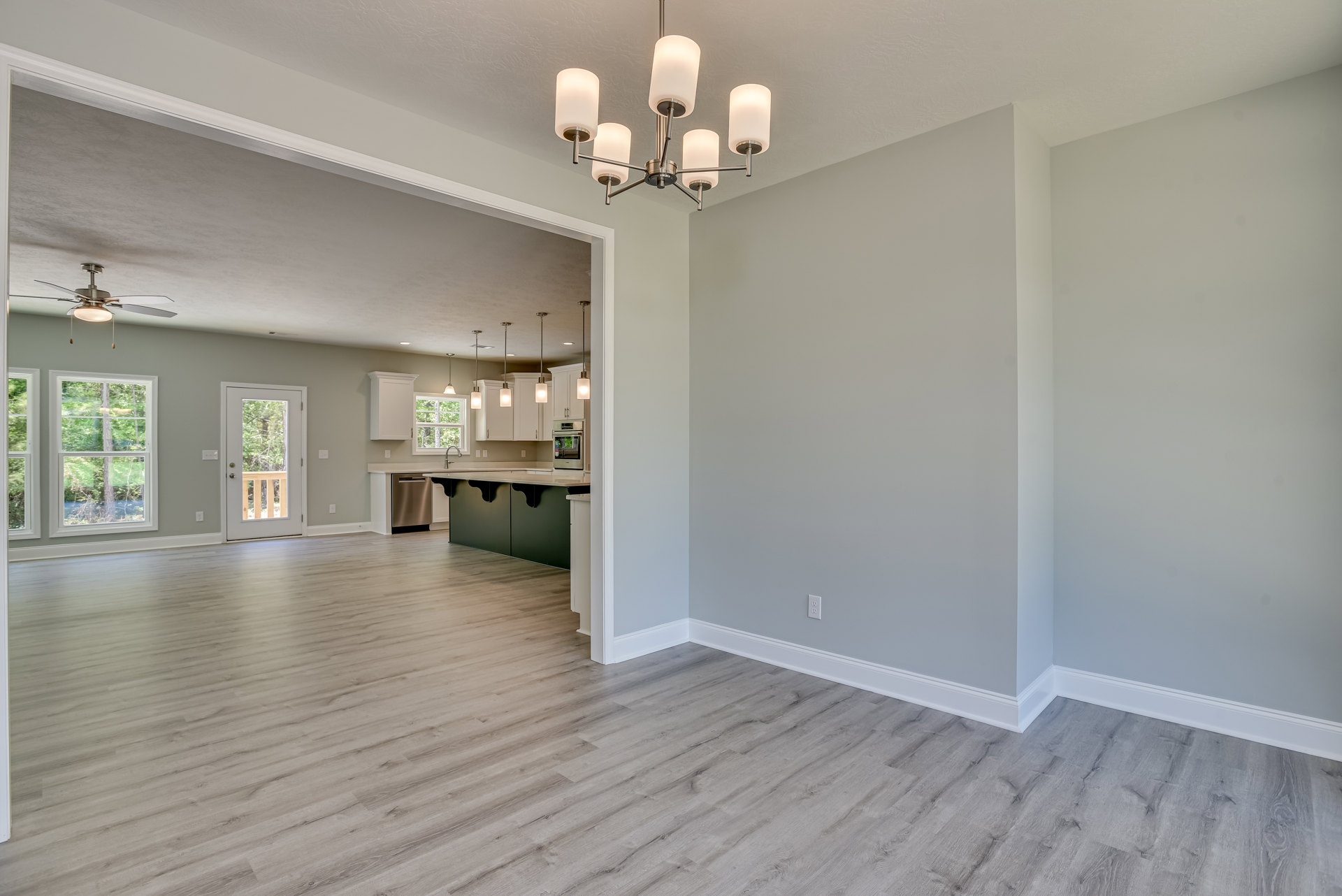 Open living room with wide plank wood flooring, white plaster walls, decorative ceiling molding, large window and glass door framing leafy trees, elegant chandelier with white