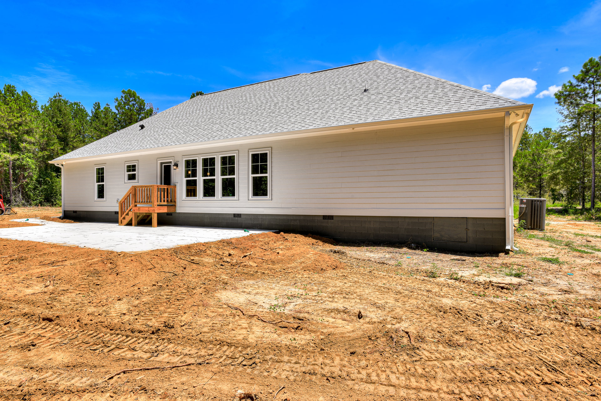 Large front porch with wooden stairs, shingle roof, white-framed windows, brick and dirt foundation, metal utility box with attached hose, surrounded by trees and sky