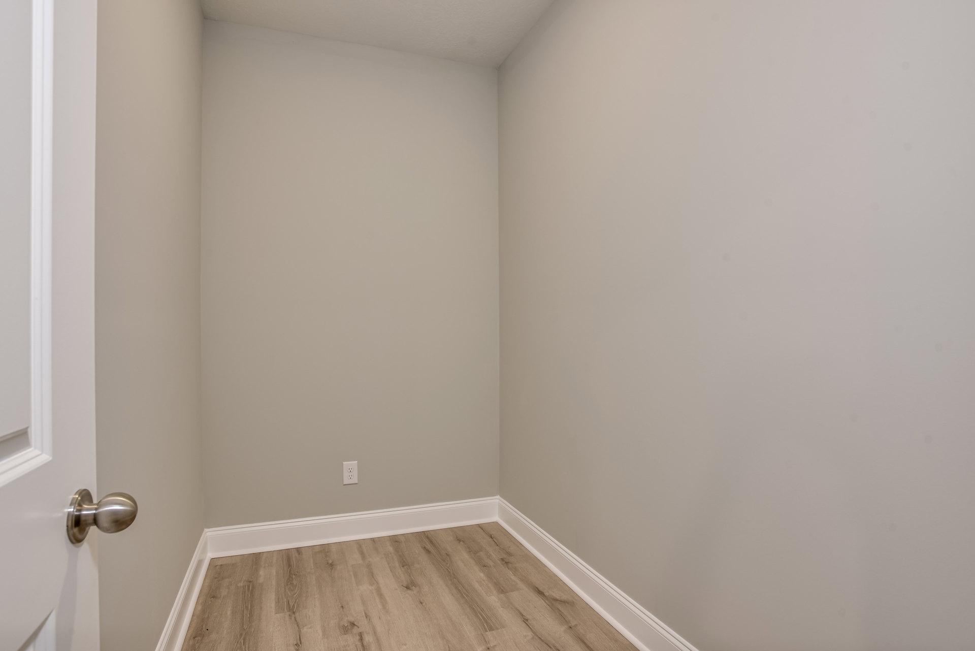 Corner of a room featuring light wood flooring, white baseboard trim, and smooth white plaster walls with a white electrical outlet and a brushed metal doorknob.