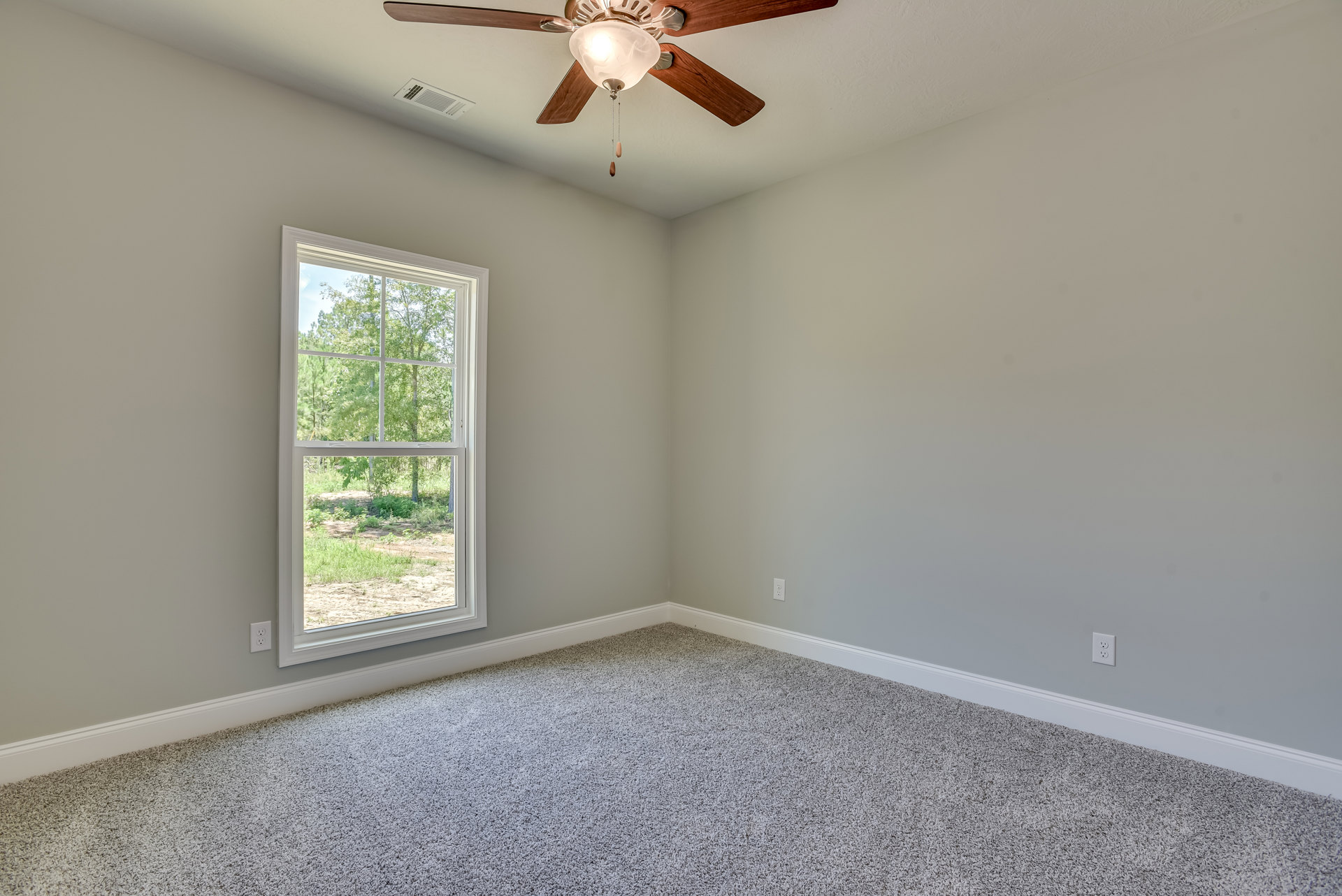 Neutral-toned carpeted room with white walls, ceiling fan with light fixture, rectangular air vent, large window showing green trees outside