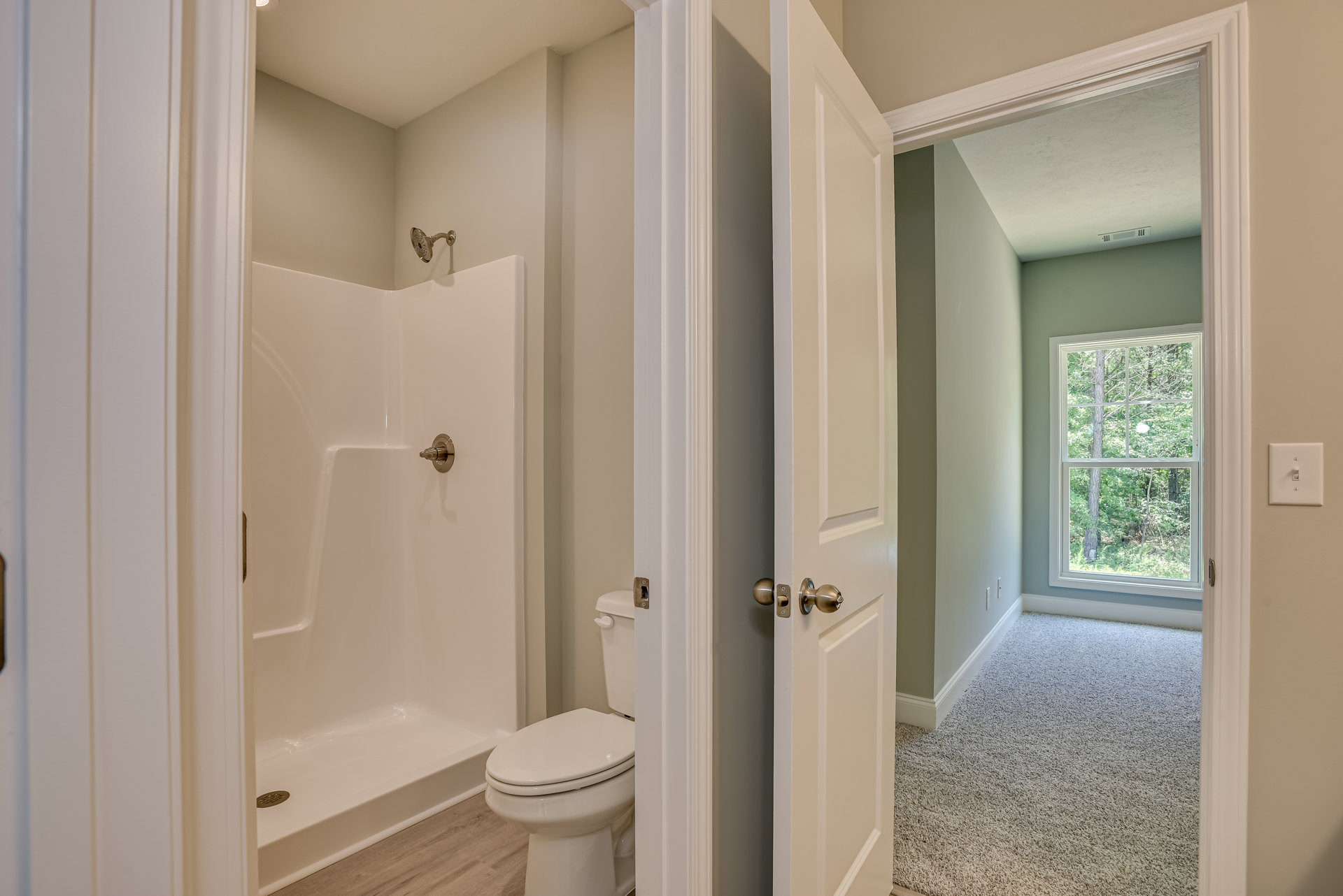 Bathroom with white toilet, glass shower enclosure, metal shower head, tile walls, window showing trees outside, metal knob, and light switch on painted wall.