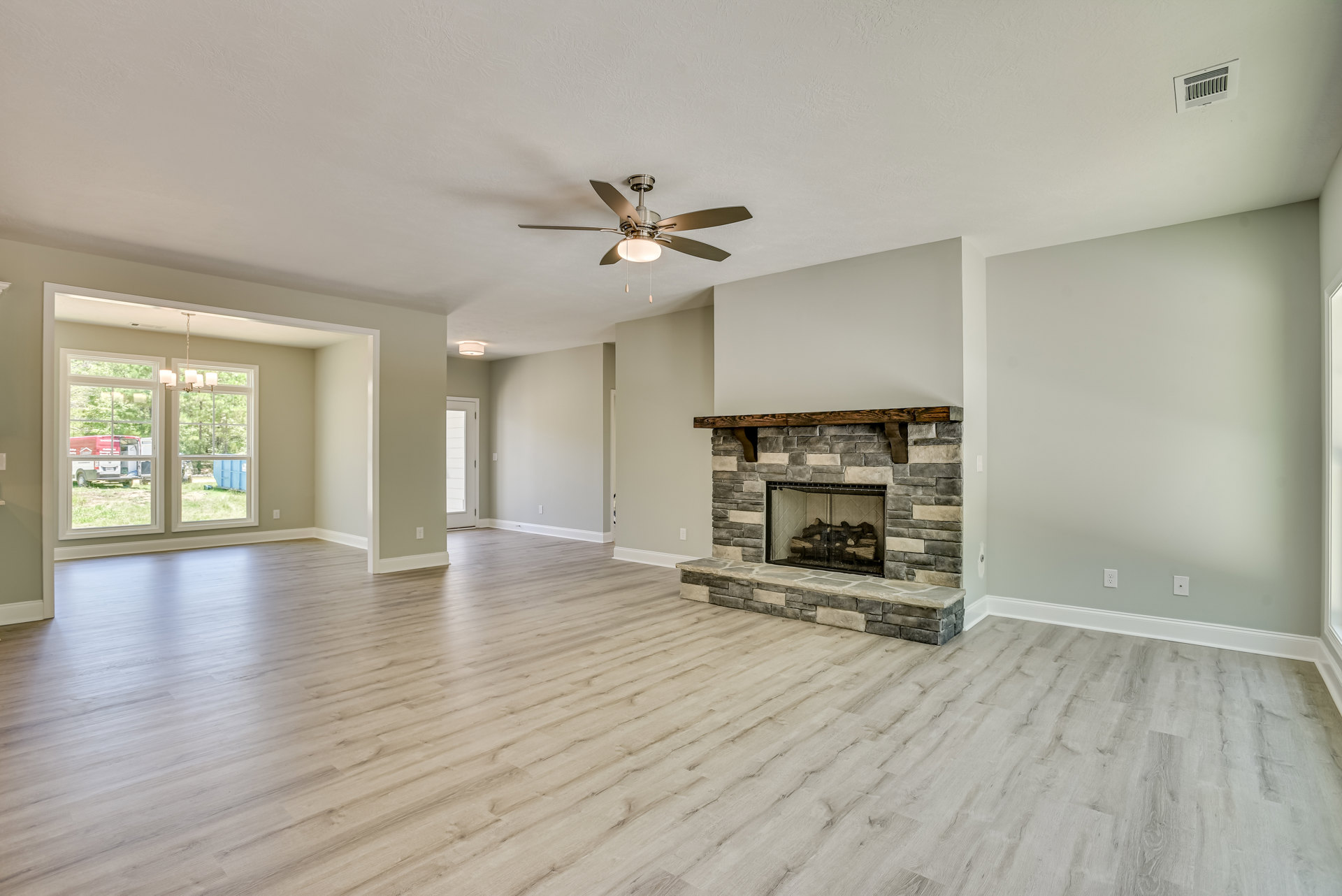Living room with wood flooring, stone fireplace filled with logs, ceiling fan with light, neutral walls, and wall vent
