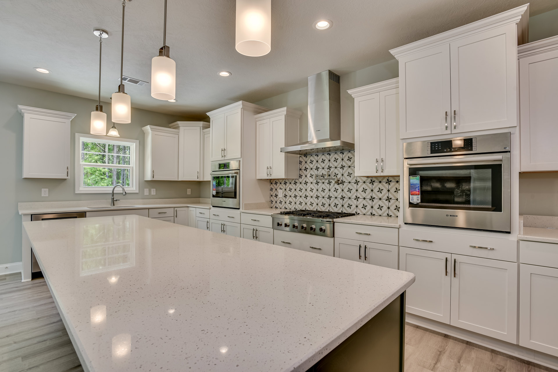 White cabinetry kitchen featuring a spacious island with white countertops, stainless steel oven and microwave, tile backsplash, sink beneath a window overlooking trees, and modern