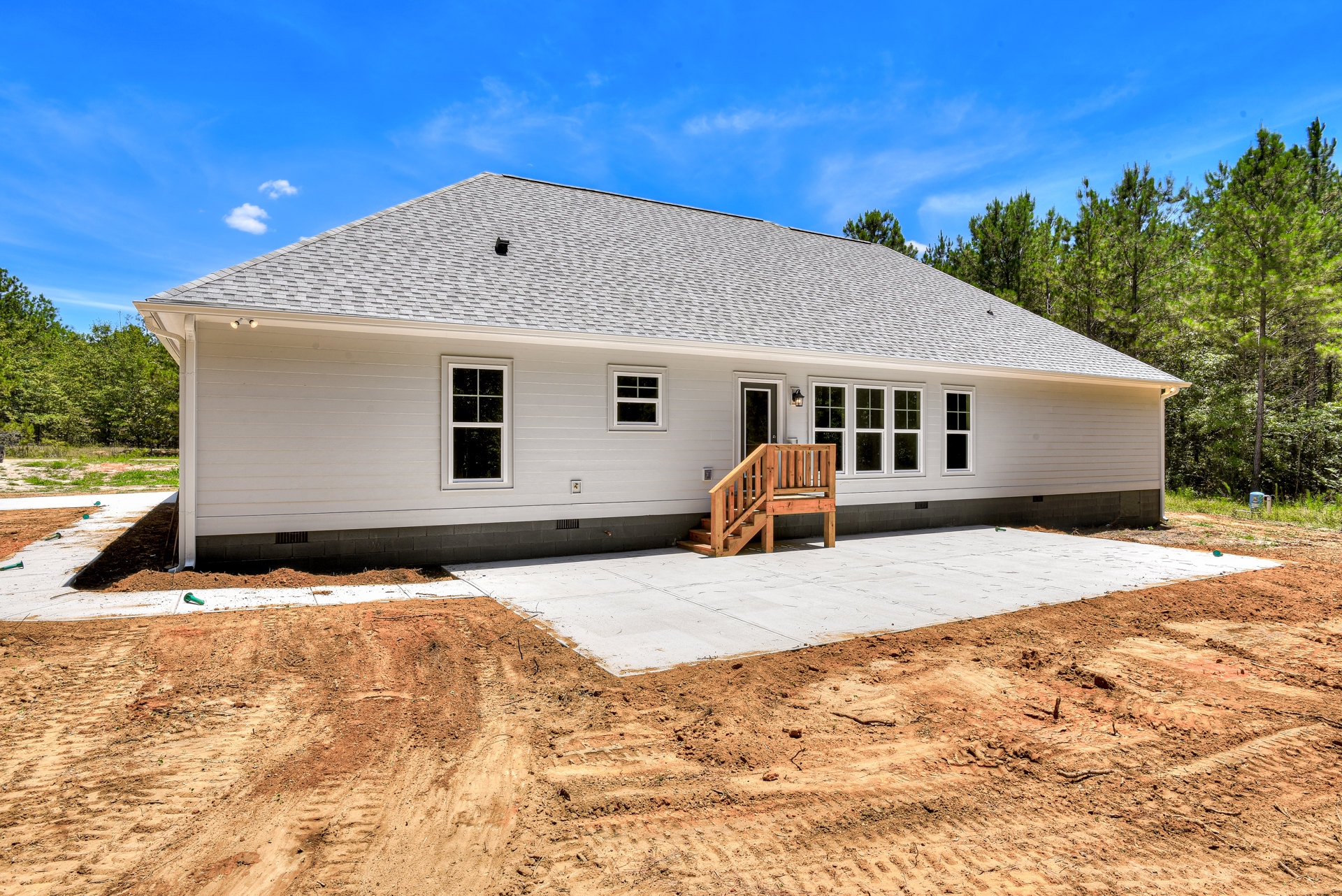 Two-story house under construction with exposed wooden staircase, white-framed windows, unfinished dirt yard, and partial roof against blue sky