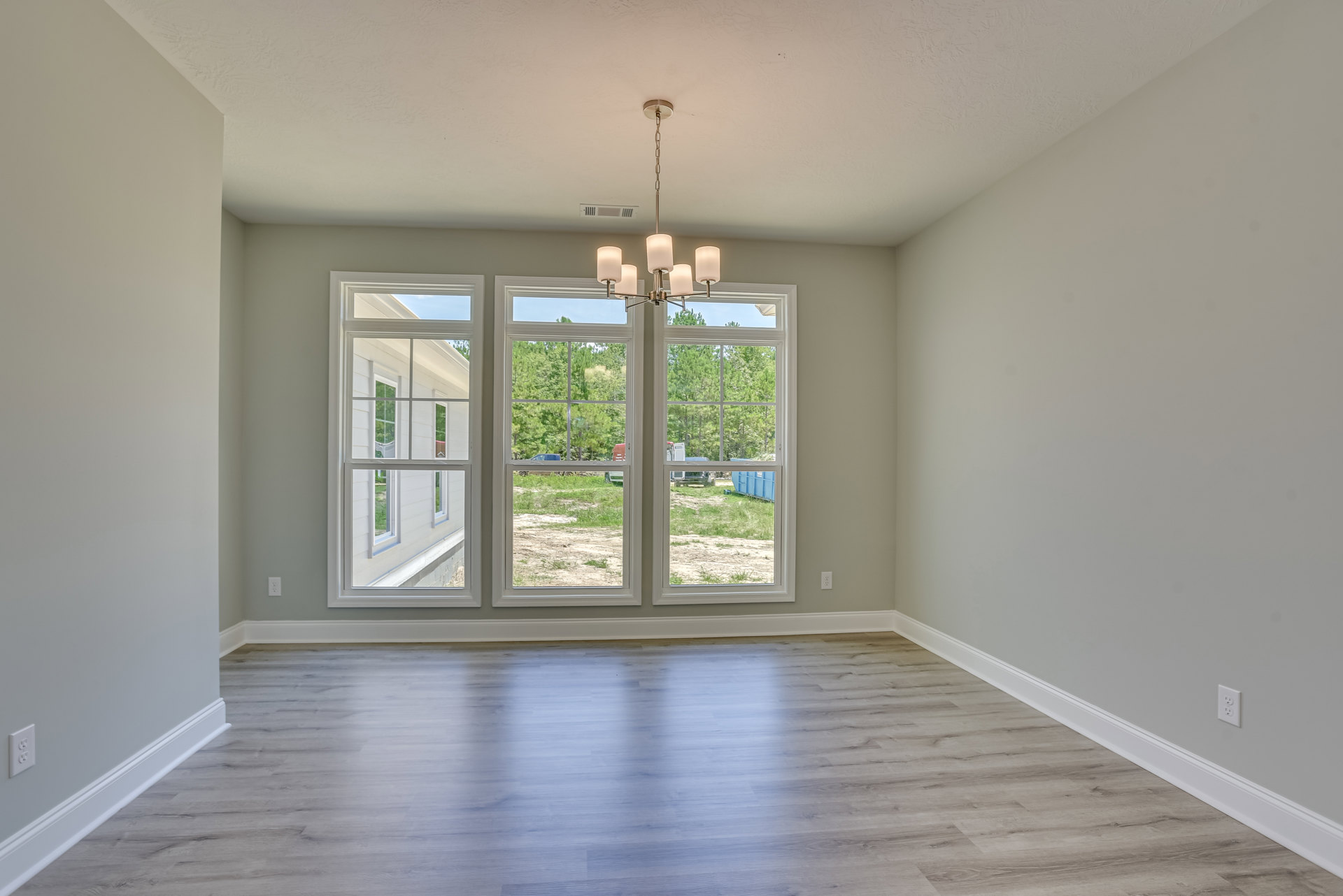 Spacious room featuring multiple large windows with white frames, natural light illuminating polished wood flooring, chandelier hanging from ceiling, white plaster walls with