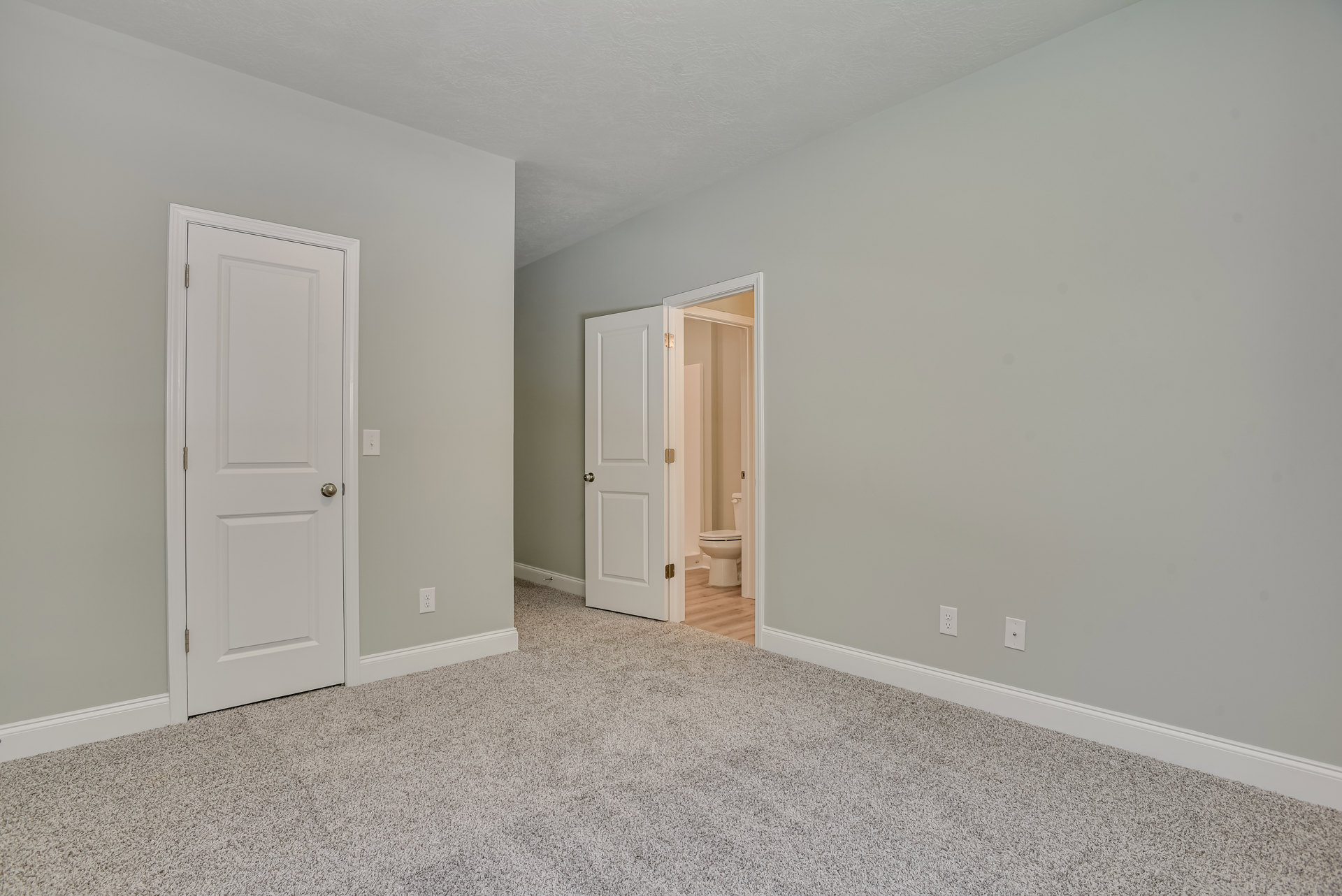 Carpeted room with white paneled doors featuring silver and gold door knobs, visible bathroom with toilet through open doorway, white walls with crown molding