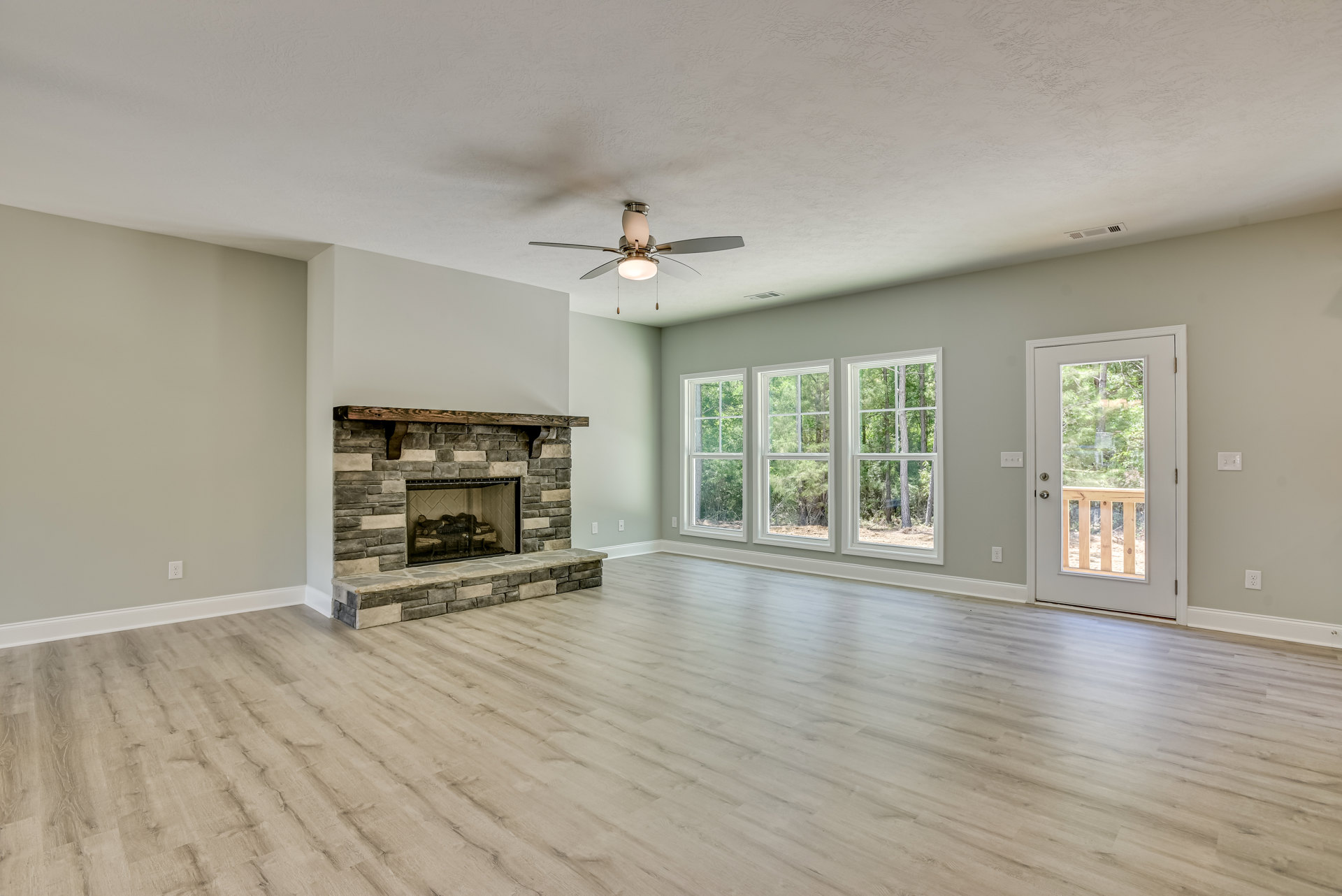 Spacious living room featuring hardwood floors, a stone fireplace with stacked logs, ceiling fan with light fixture, glass-paneled door, and a row of windows overlooking leafy