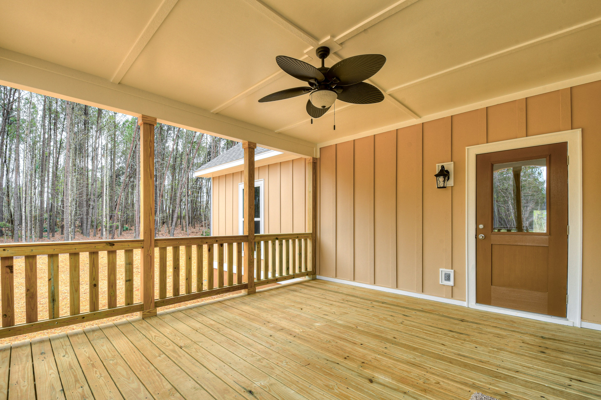 Covered porch with wood plank flooring, ceiling fan with light, glass-paneled door, wooden railing, and forest view in the background