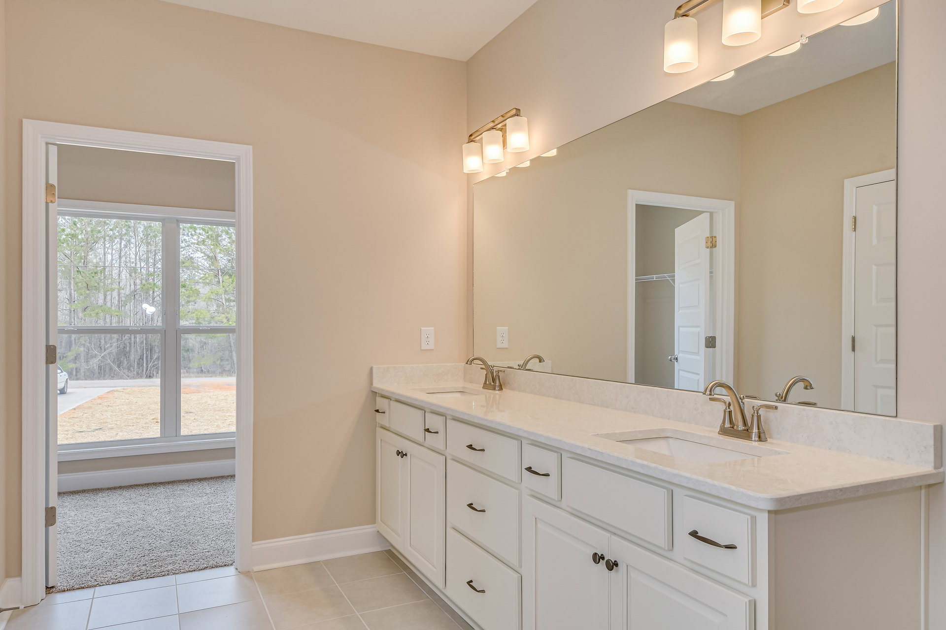 Bathroom with expansive mirror above double sinks, white cabinetry and countertops, window overlooking trees and parked car, tiled floor, modern plumbing fixtures, and sleek drawer