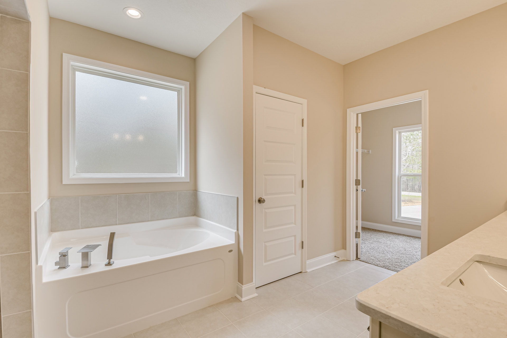 Bathroom with white square sink on a countertop, freestanding bathtub, frosted glass window, white door with silver doorknob, light tile walls and floor.