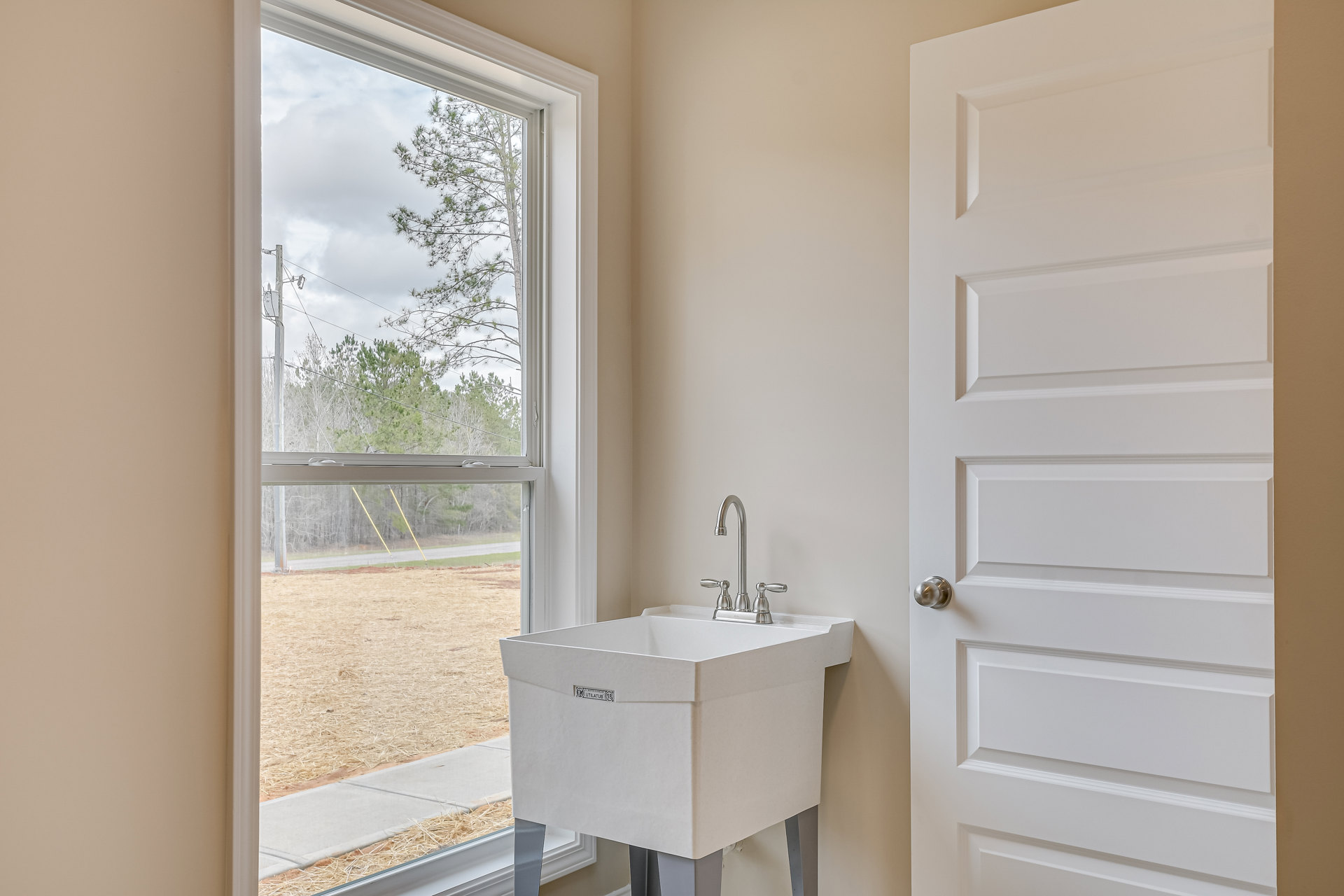 White ceramic sink with chrome faucet beneath a window overlooking trees and power lines, surrounded by light-colored tile walls; white door with silver knob visible to the side.