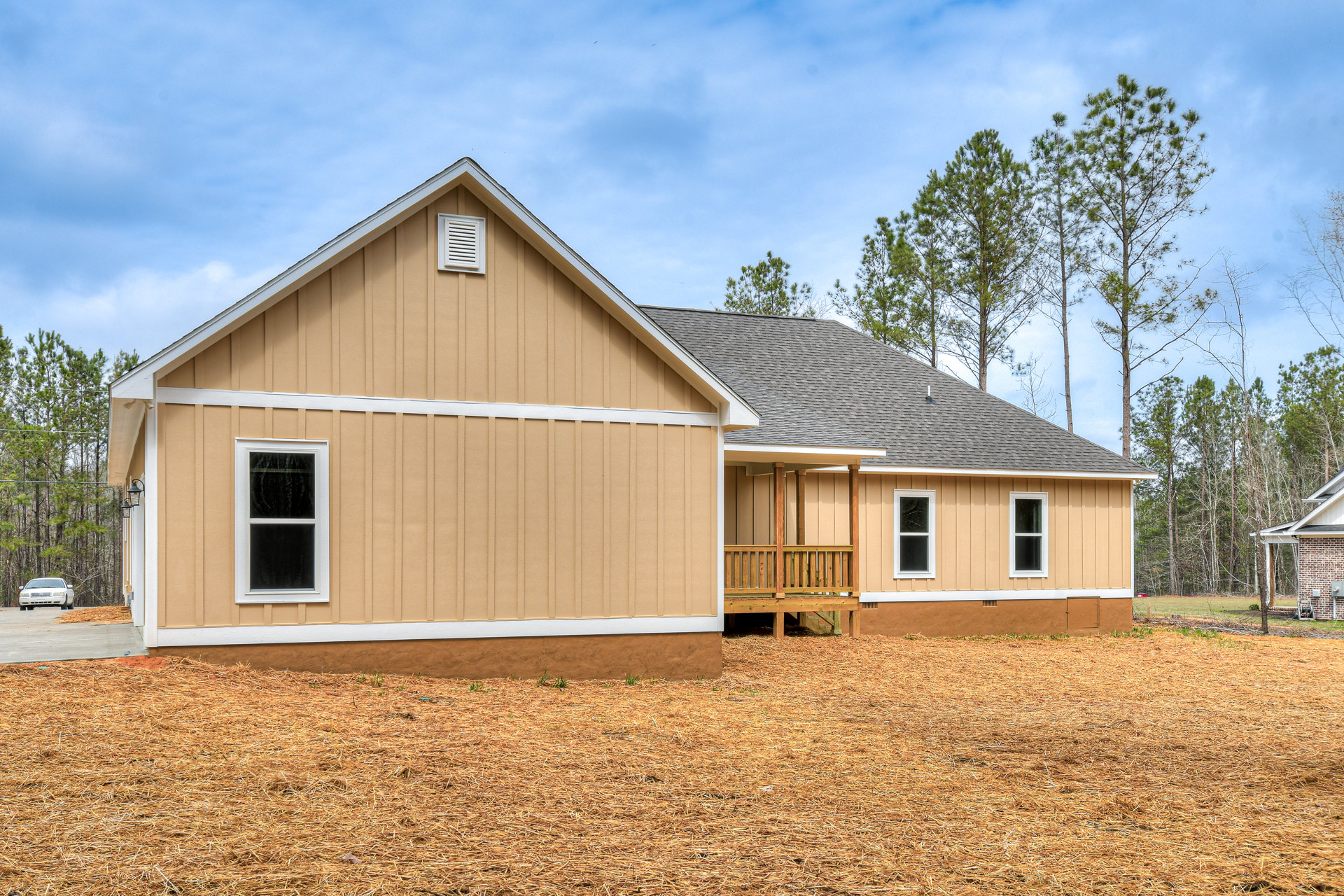 Two-story house with covered front porch, wooden deck railing, white vent on brown siding, multiple windows including one with broken glass, grassy yard, mature trees, and partly
