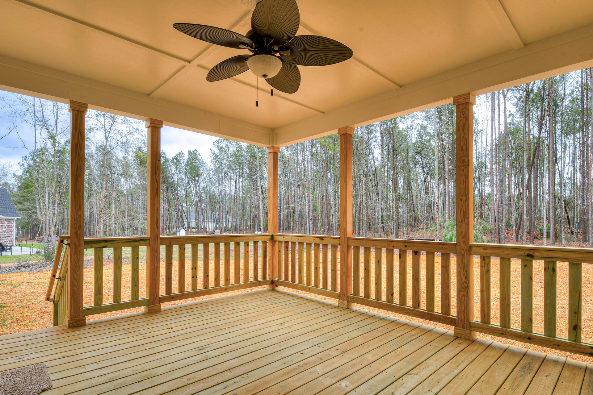 Covered porch with wood decking, ceiling fan with light fixture, white railing, brick exterior wall, and leafy trees in the background