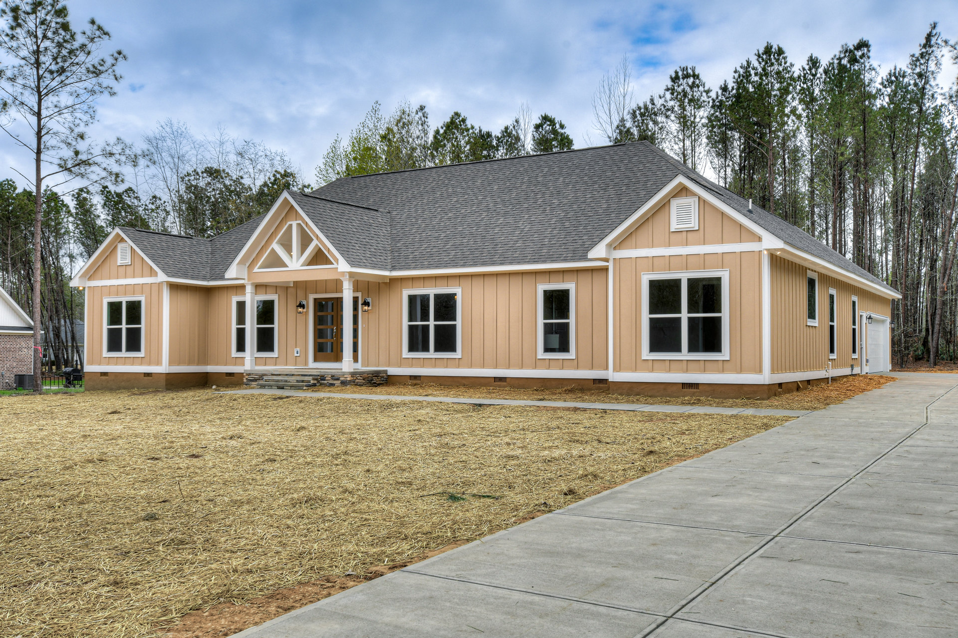 Two-story house with white siding, covered front porch, paved driveway, large windows with white frames, surrounded by mature trees and landscaped lawn under a partly cloudy sky