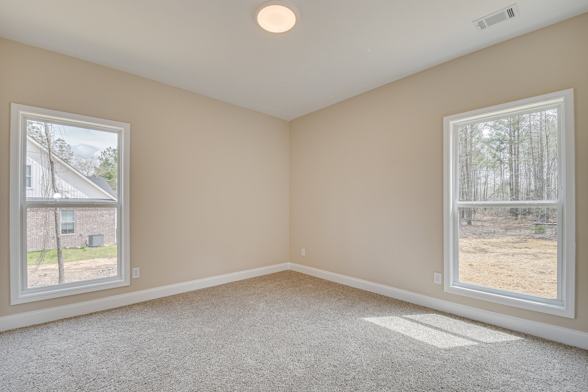 Carpeted room with two large windows, white walls, ceiling light fixture, view of trees and neighboring house through windows