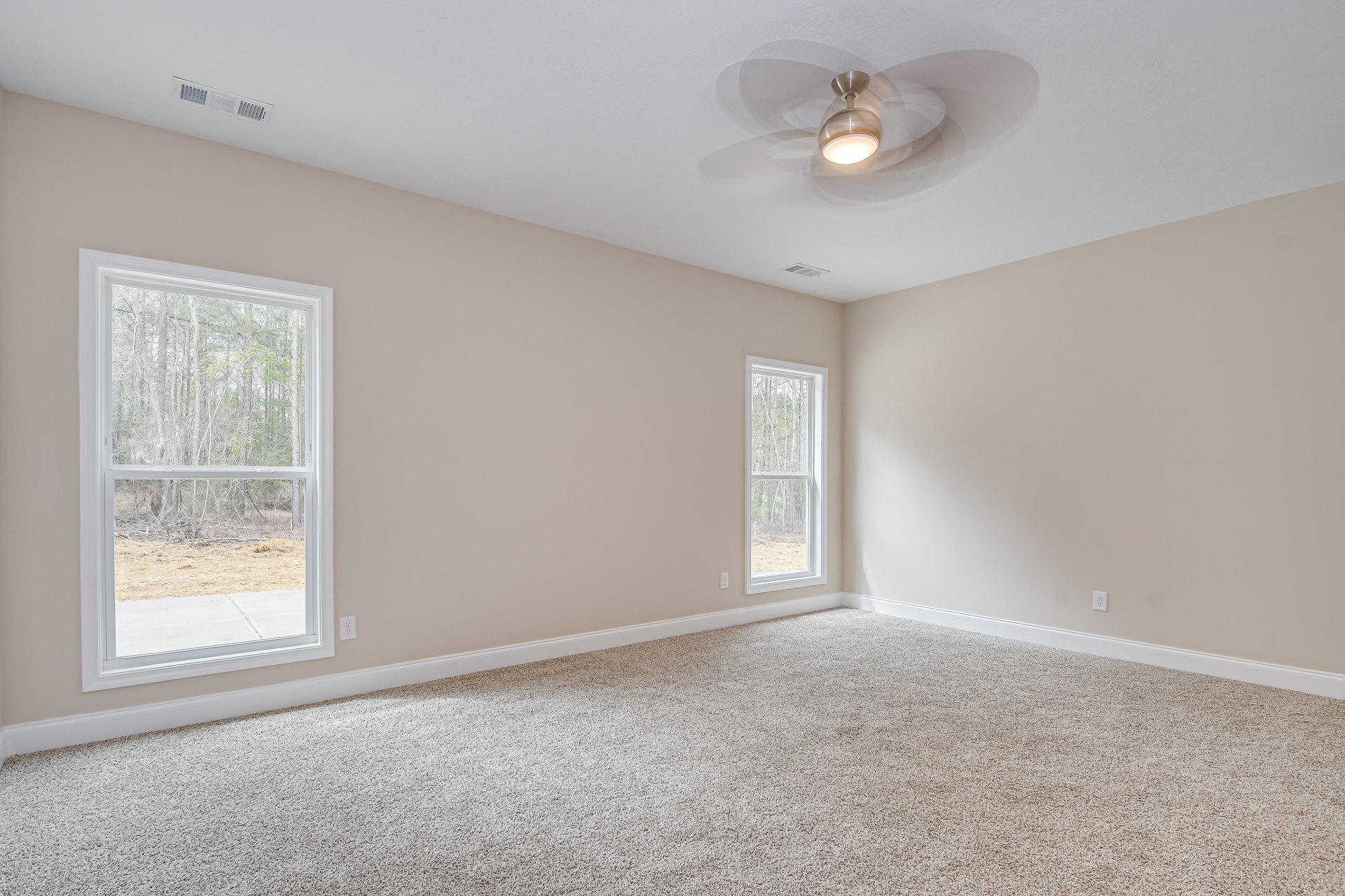 Carpeted bedroom with white walls, ceiling fan and light fixture, large windows overlooking forested landscape, plaster ceiling with air vent and crown molding.