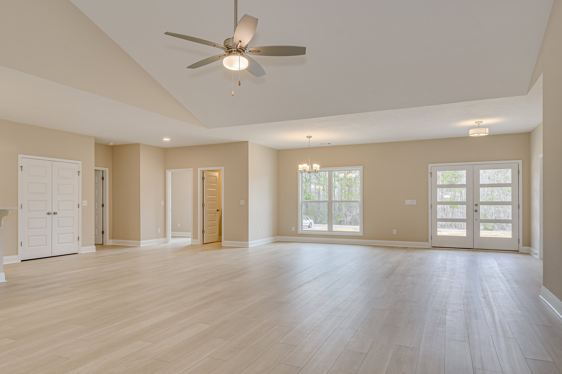 Spacious room with wood flooring, ceiling fan with light fixture, white double doors featuring glass panes and silver handles, window showing parked car outside, white walls with