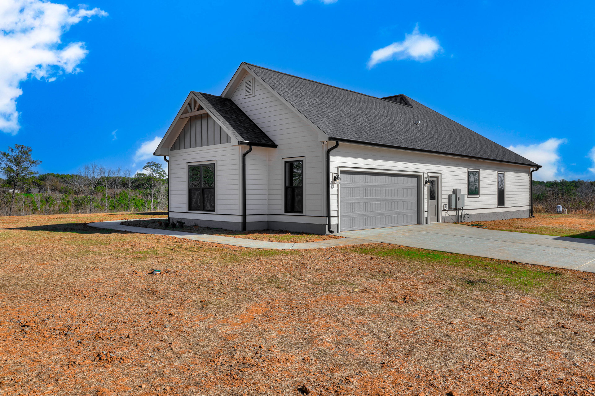 Two-story home with gray siding, white trim, attached garage, concrete driveway, manicured lawn, and large windows reflecting surrounding trees under a clear blue sky