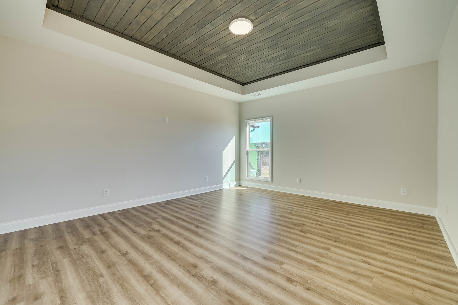 Open room with wood plank flooring and matching wood ceiling, large window framing leafy trees, plaster walls, ceiling-mounted light fixture illuminating space
