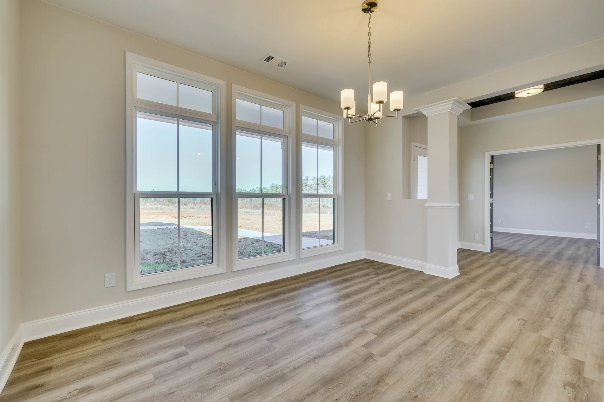 Room with light wood flooring, white-framed windows, open door, and view of trees and dirt field outside