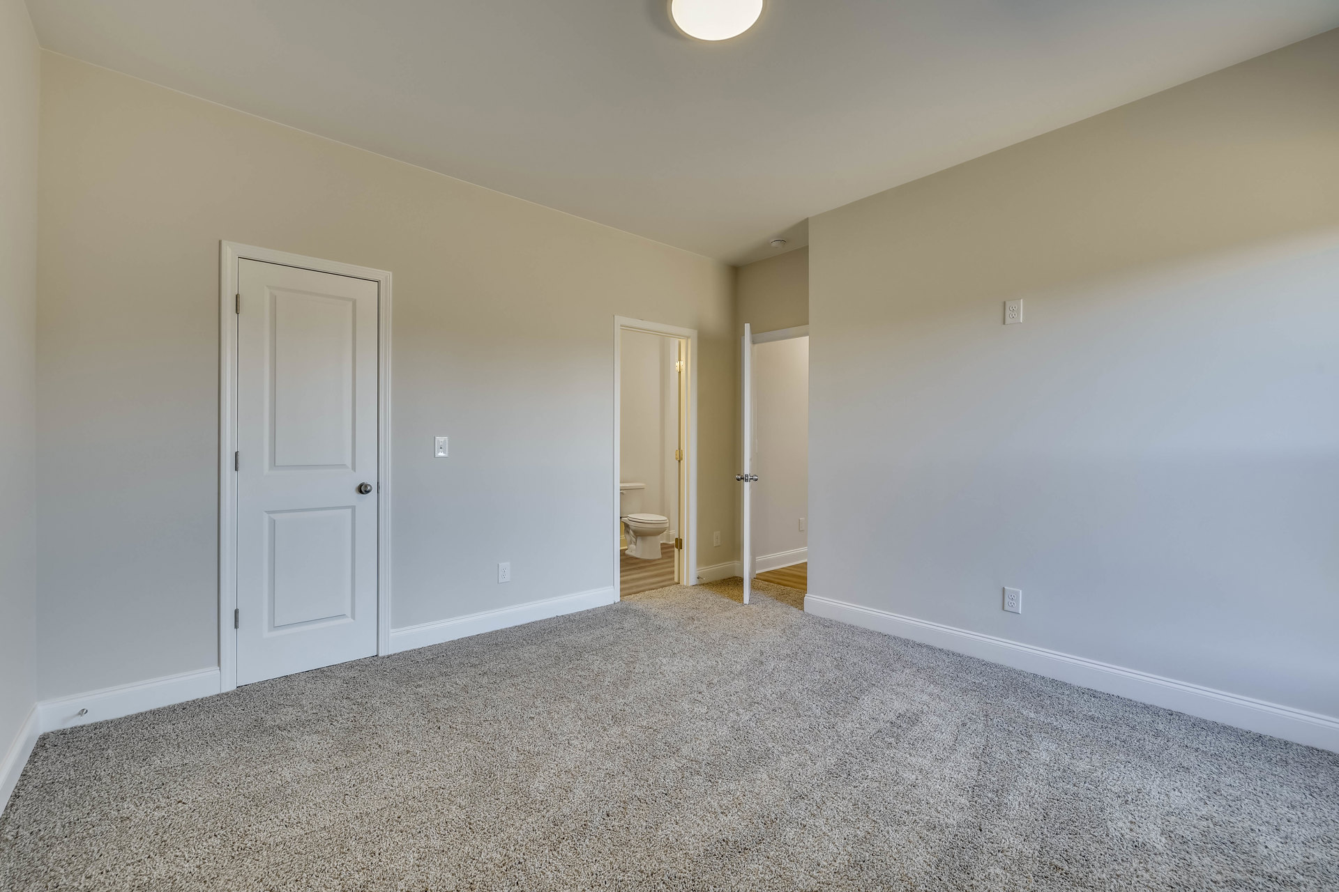 Carpeted room with white walls, white square-framed door with silver knob, desk, and visible bathroom with toilet